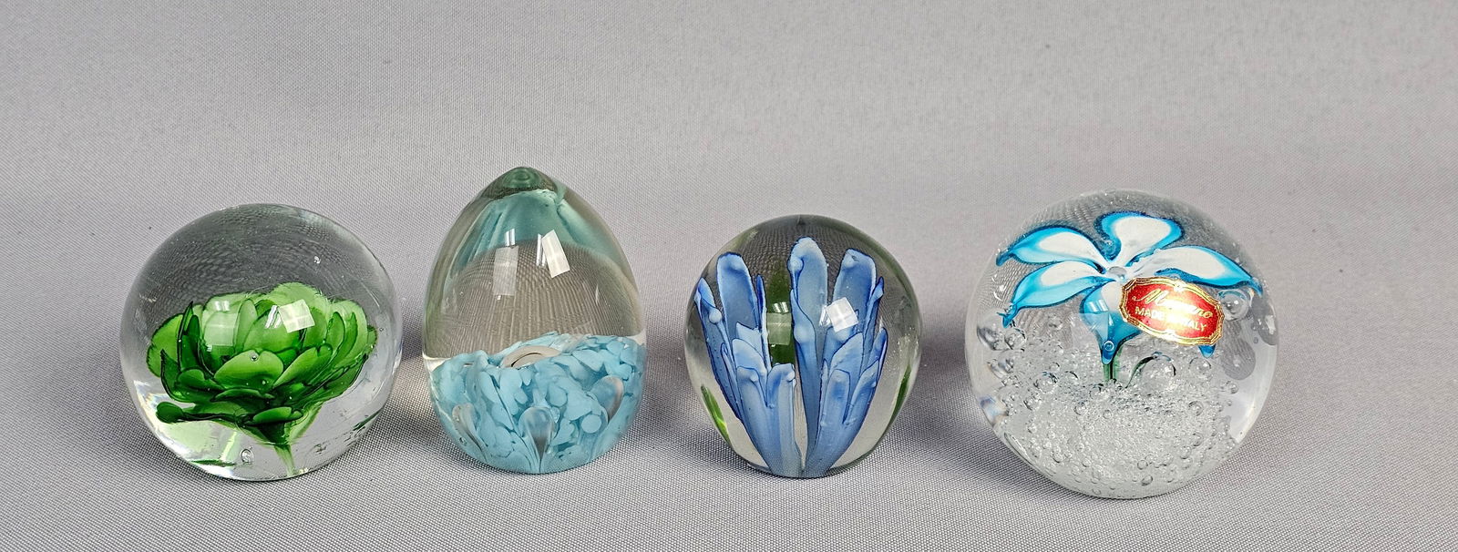 A Group of Four Decorative Glass Paperweights Including Murano: A group of four contemporary decorative glass paperweights, each crafted from clear glass encapsulating various internal colored glass designs. The final spherical paperweight showcases a prominent bl