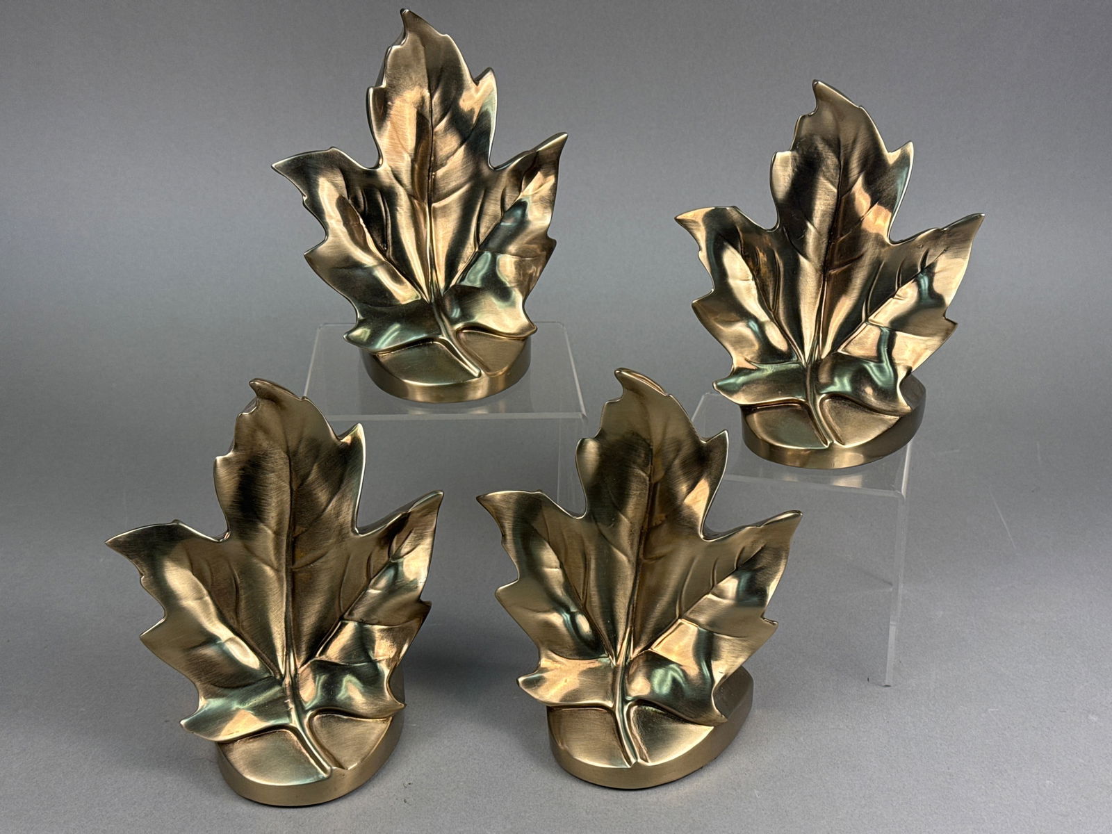 A Set of Four Mid-20th Century PM Craftsman Brass Maple Leaf Bookends: A set of four Mid-20th Century PM Craftsman Brass Maple Leaf Bookends. Each bookend is crafted from a brass or bronze-toned metal with a reflective, lacquered finish, depicting a maple-like leaf mount