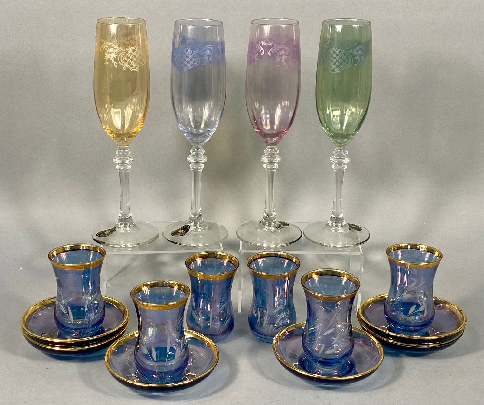 A Set of Four Cristalleria Fumo Multicolored Flute Goblets and a Set of Six Iridescent Purple-Blue: A set of four Cristalleria Fumo multicolored flute goblets, each featuring a clear stemmed base with a colored bowl (yellow, light blue, pink, and green) adorned with an etched repeating scrollwork pa