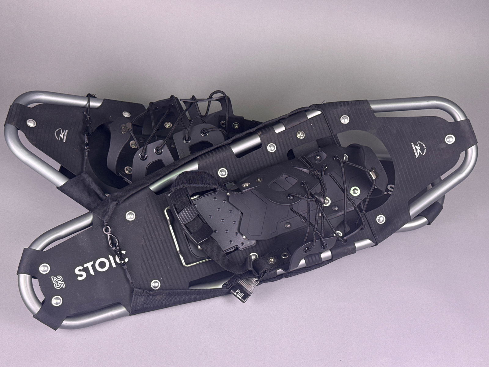 A Pair of Stoic 25in Snowshoes: A pair of Stoic 25in snowshoes. Primarily constructed with a gray aluminum frame, black fabric decking, and black plastic bindings featuring metal crampons and hardware. Visible markings include 'STOI