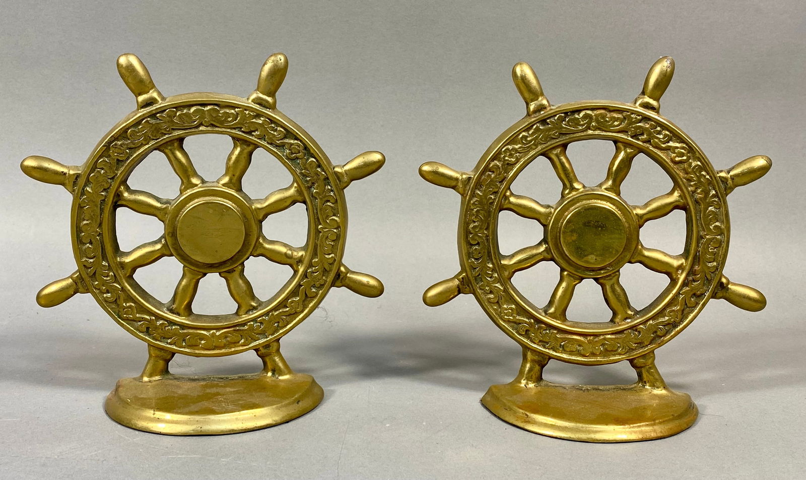 A Pair of Vintage Brass Nautical Ship's Wheel Bookends: A pair of vintage brass nautical ship's wheel bookends. Each solid brass decorative item is shaped as a six-spoked ship's wheel with six handles. The outer ring of the wheel features a continuous, orn