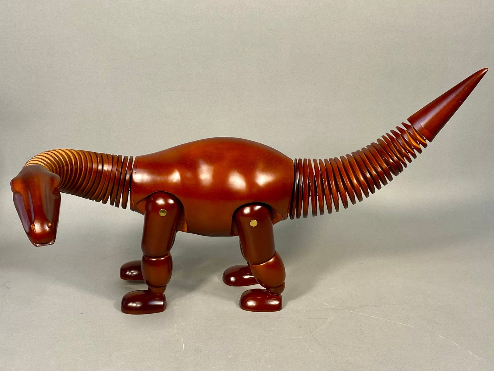 Wooden Brontosaurus Dinosaur Mannequin with Flexible Neck and Tail: A fully articulated wooden figure, crafted from a reddish-brown hardwood with a high-gloss, polished finish. It features a smooth, solid-looking torso, articulated legs with brass-toned metal pin join