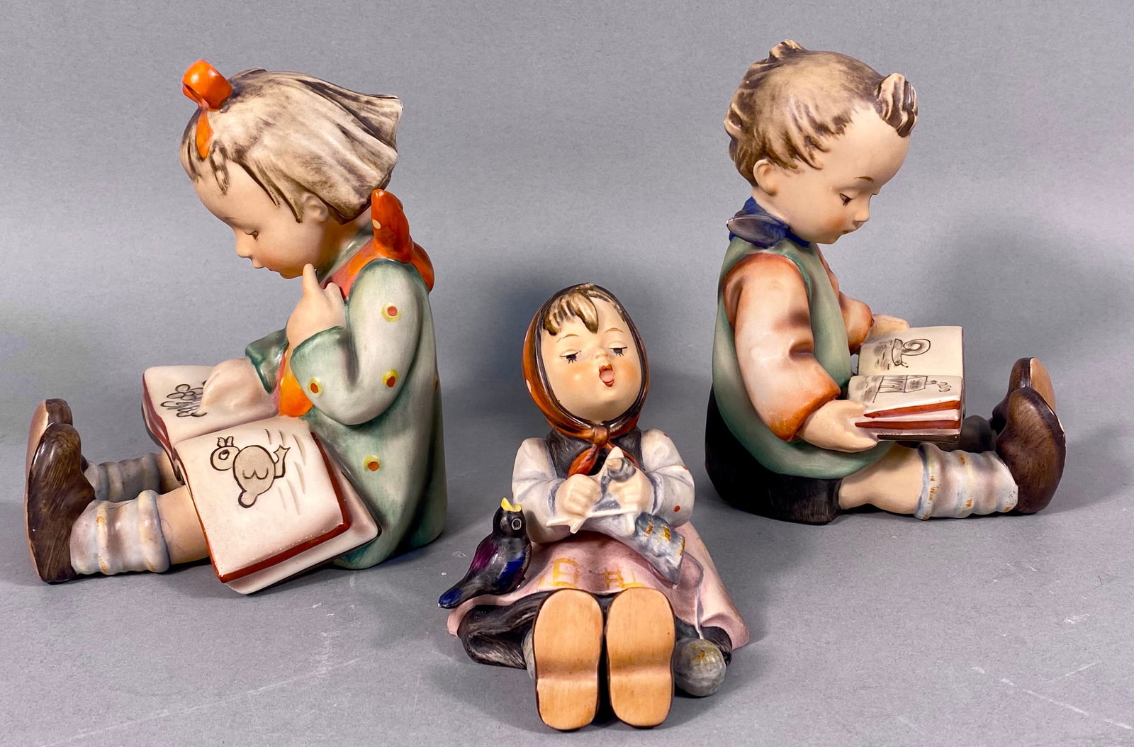 Three M.I. Hummel Goebel Figurines, including 'Happy Pastime' #69: A group of three hand-painted ceramic figurines by M.I. Hummel Goebel. The lot includes an M.I. Hummel Goebel 'Happy Pastime' figurine, model number 69, depicting a girl seated, knitting, with a small
