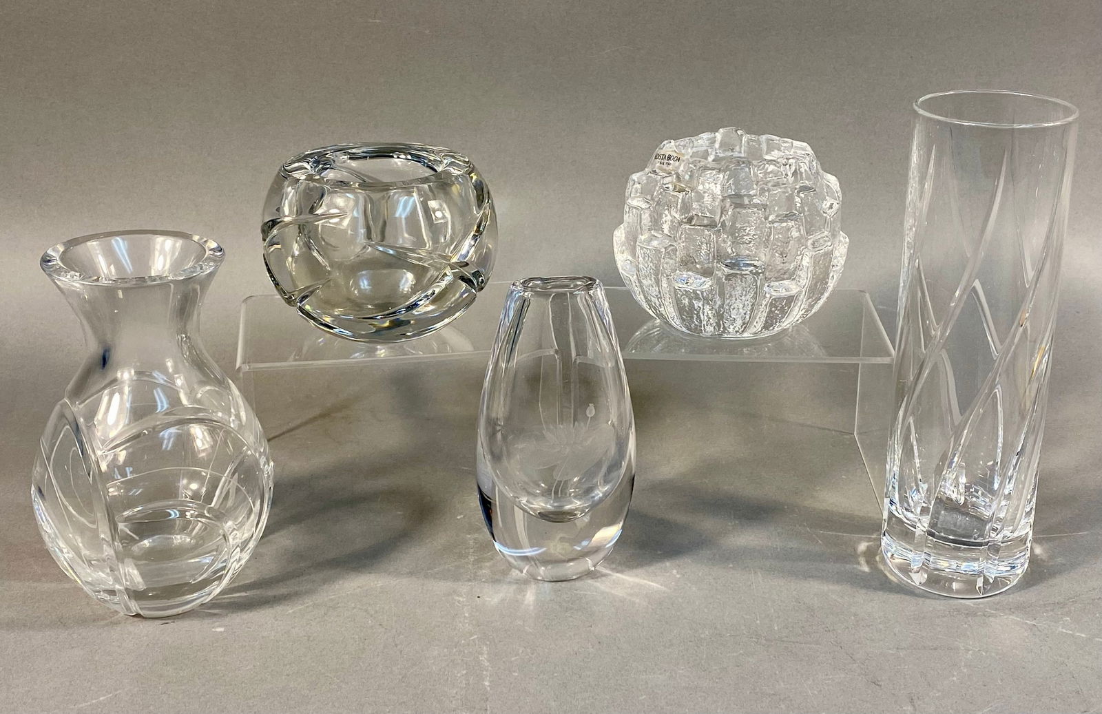 A Group of Five Assorted Glass and Crystal Vases and Votives, including Kosta Boda, Hoya, Miller: A collection of five clear glass and crystal decorative objects, comprising three distinct vases and two spherical votive or small vase forms. The items exhibit a range of contemporary styles, featuri