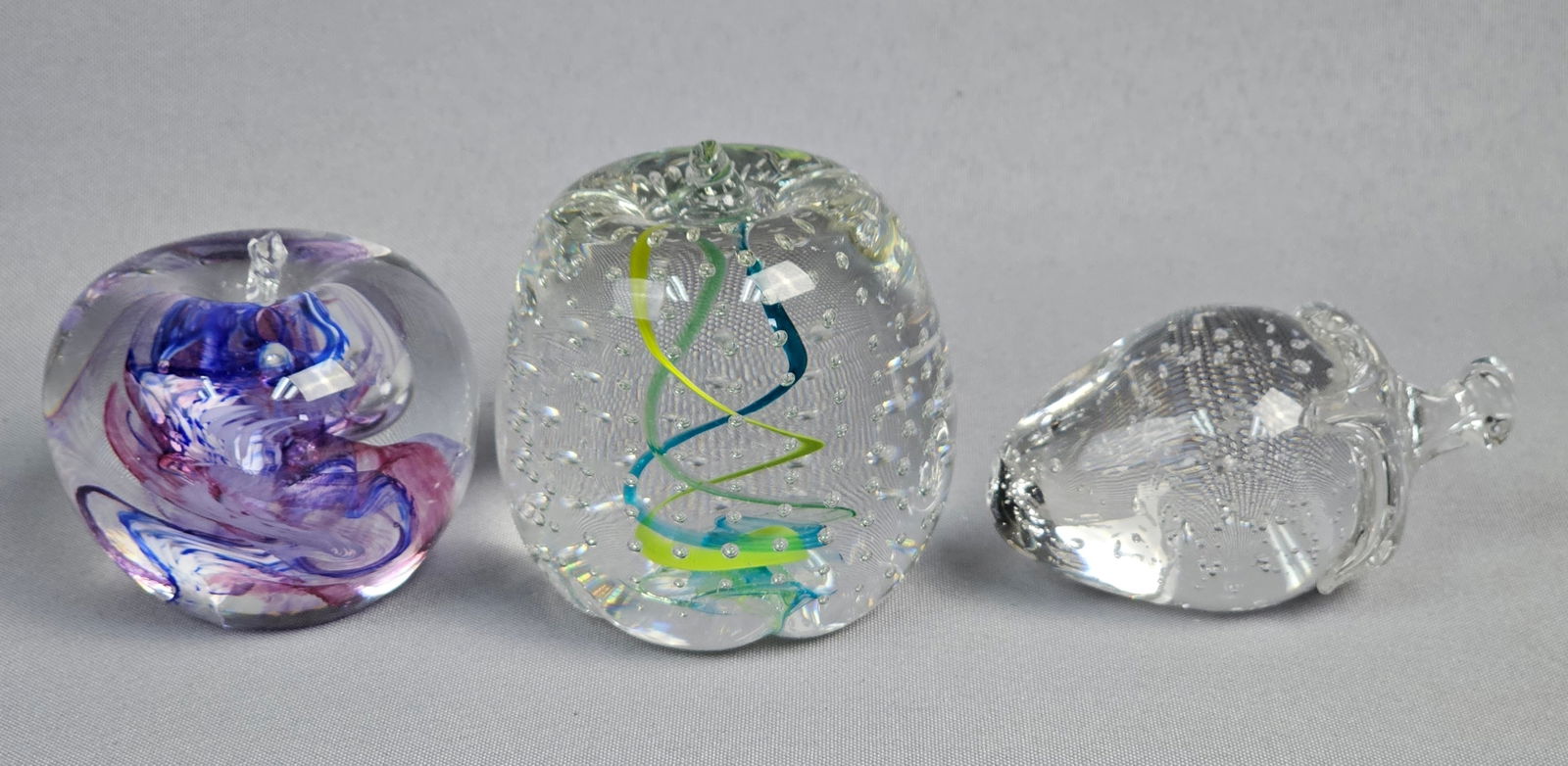 Three Contemporary Art Glass Apple and Strawberry Paperweights: This lot comprises three contemporary art glass decorative objects. The leftmost piece is a clear glass apple-shaped paperweight, featuring internal abstract swirls in shades of purple, blue, and pink