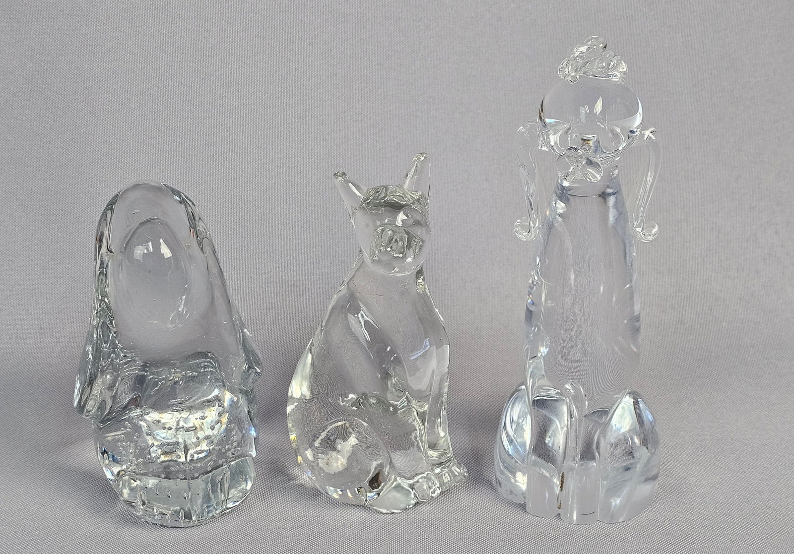 Three Clear Art Glass Figurines Paperweights: A group of three clear, colorless glass figurines, exhibiting a modernist or contemporary aesthetic. No discernible maker's marks or textual inscriptions are visible on any piece. Pieces measure 4.5-6