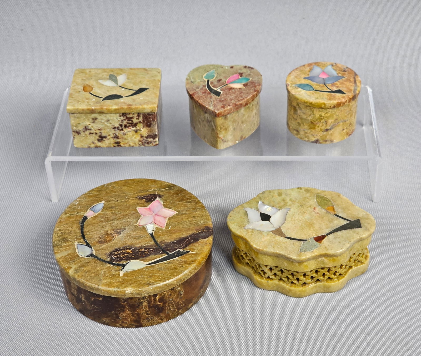 A Collection of Five Soapstone Inlaid Floral Trinket Boxes and One Candle Holder, Made in India: A collection of five lidded decorative boxes and one candle holder, crafted from natural stone, likely soapstone or marble, displaying variegated tones of tan, brown, and green. Each lidded box and th