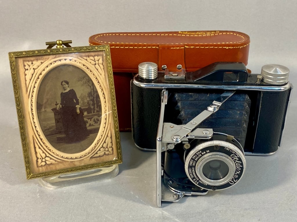 A Foldex 20 Folding Camera with Case and a Victori: The lot comprises a vintage Foldex 20 folding camera with its accompanying brown leather case and a Victorian tintype portrait set within an ornate frame. The camera, manufactured by PFH-F.P. Corp. of