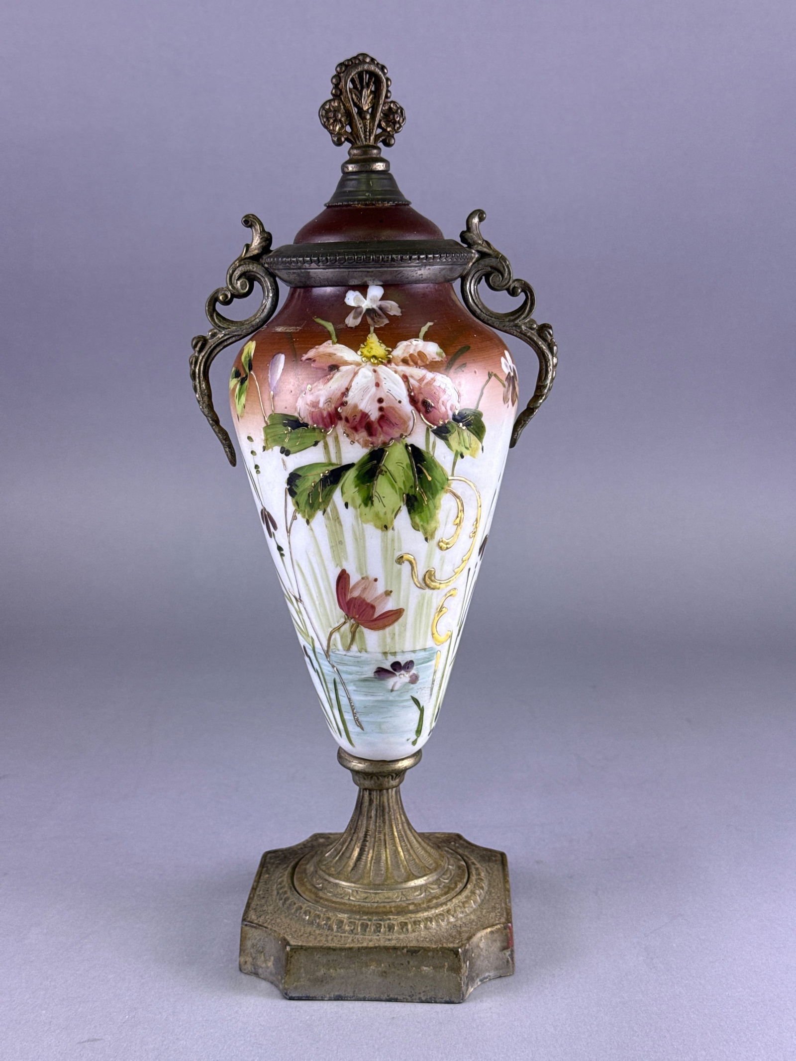 A Victorian Bronze-Mounted Polychrome Enamel Decorated Opal Glass Covered Urn: A single covered urn (vase couvert) dating to the late 19th century, constructed of opaque opal glass fitted with elaborate patinated brass or bronze metal mounts. The tapering glass body is decorated