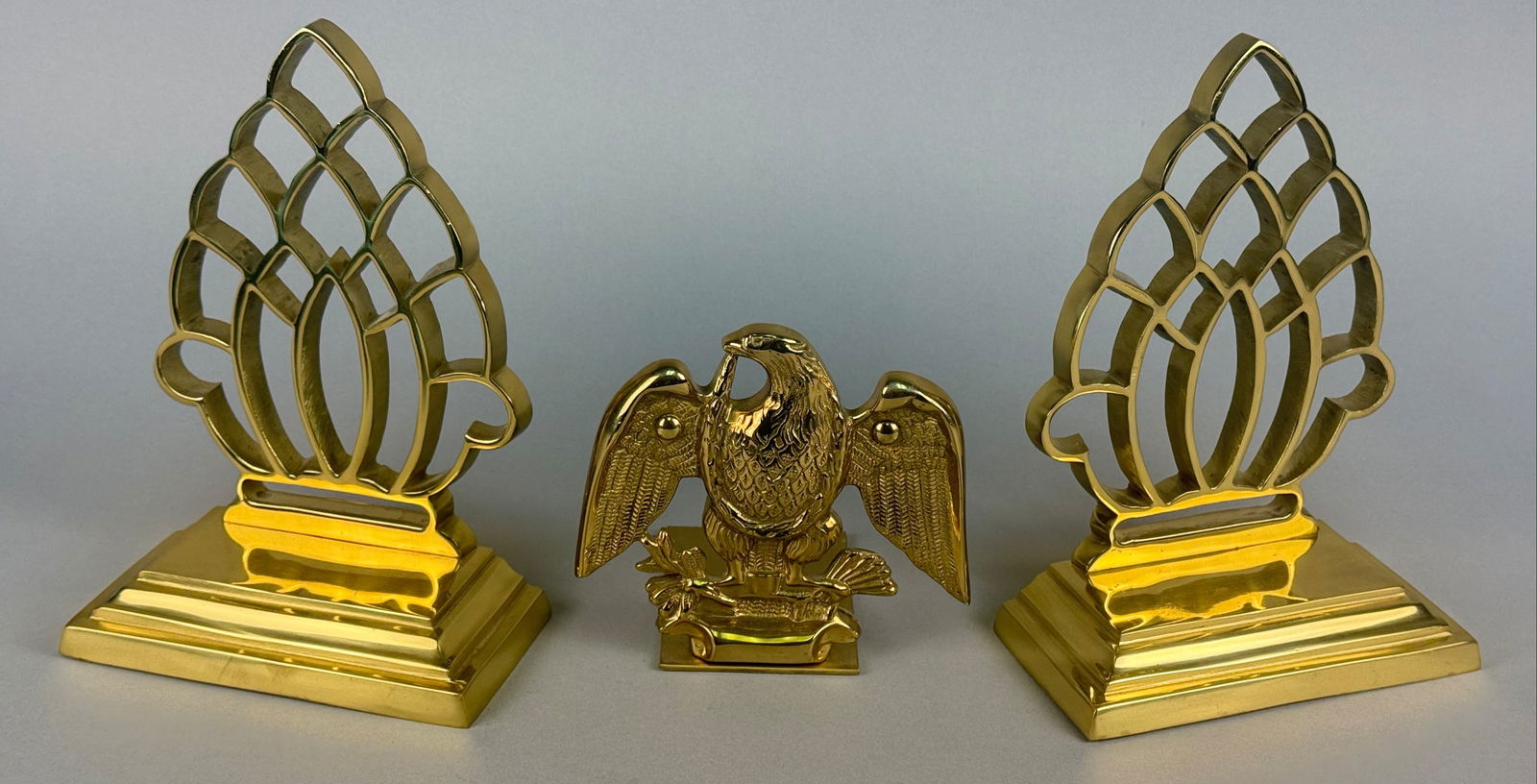 A Trio of Decorative Crafts Inc. Federal Revival Brass-Tone Bookends and Eagle Figure: A trio of decorative items comprising a pair of matching bookends and a central figure, manufactured by Decorative Crafts, Inc. The pieces are constructed from polished brass-tone metal, likely coated