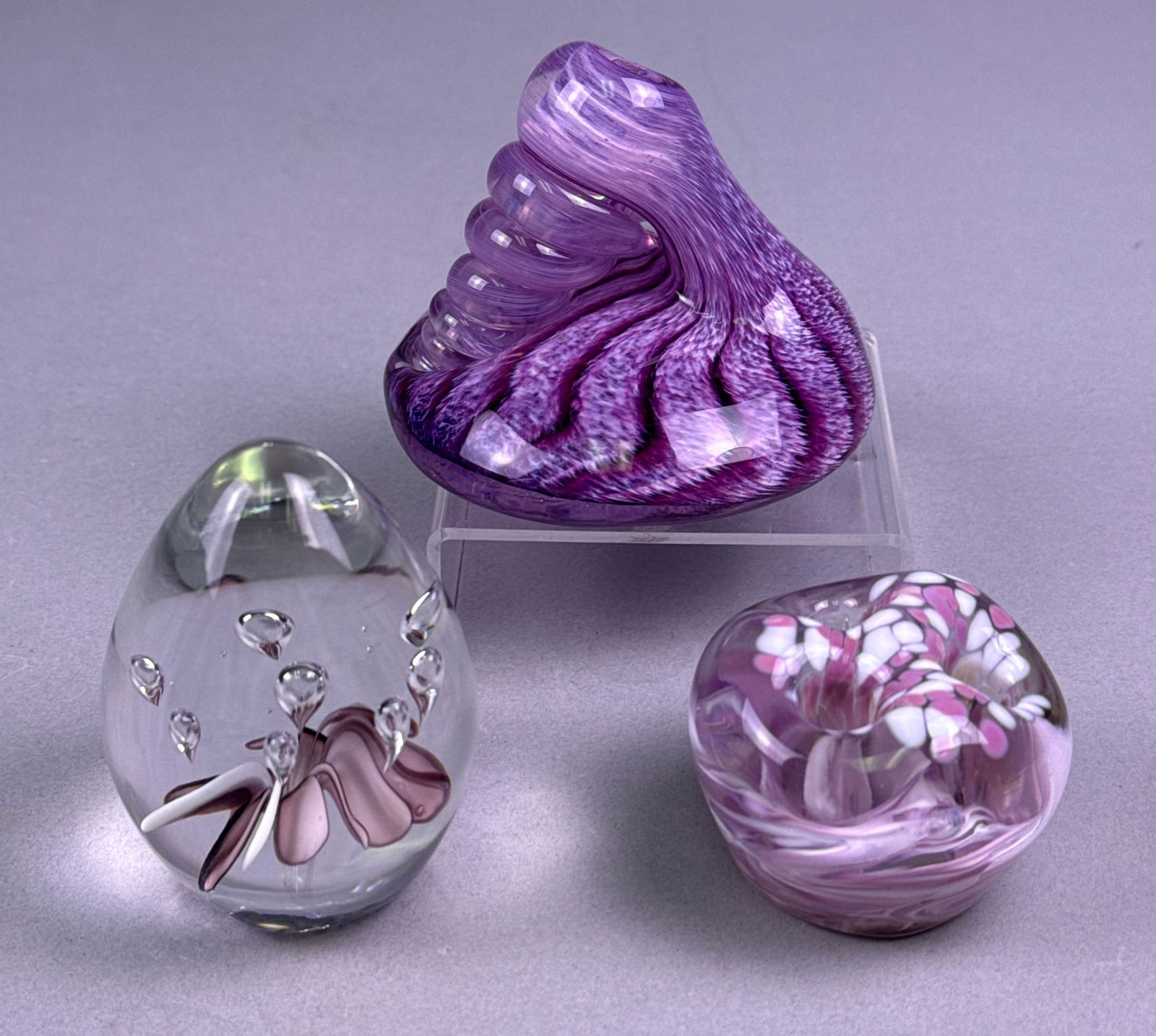 A Trio of Modern Studio Art Glass Paperweights and Sculpture in Purple and Pink Tones: A trio of modern studio art glass decorative objects, consisting of two paperweights and one conical sculpture, primarily featuring purple, pink, and clear glass. The items are composed of solid, hand
