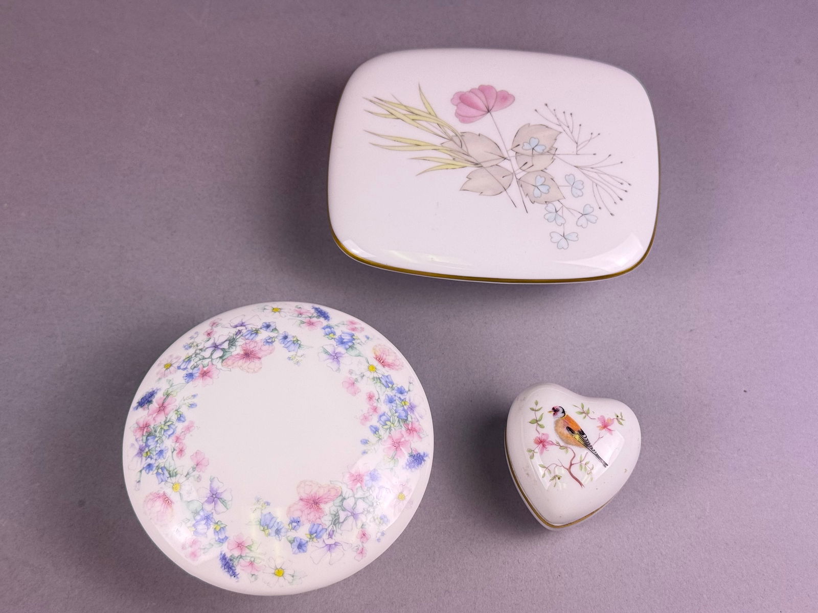 A Trio of Porcelain and Bone China Lidded Trinket Boxes: A collection of three lidded decorative boxes, varying in material and origin, featuring polychrome floral and avian designs. The grouping includes one rectangular porcelain box marked Kaiser Germany,