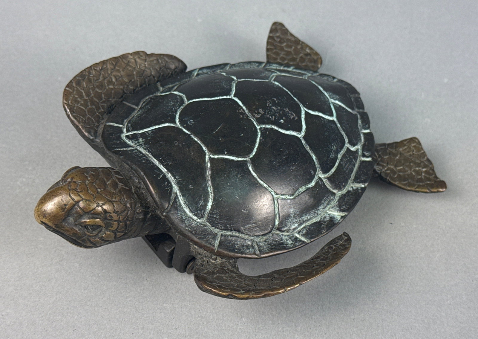 A Cast Metal Sea Turtle Door Knocker: A cast metal sea turtle door knocker, depicted in a naturalistic style. The object functions as a door knocker, featuring a hinged mechanism on the underside with mounting holes for attachment. The tu