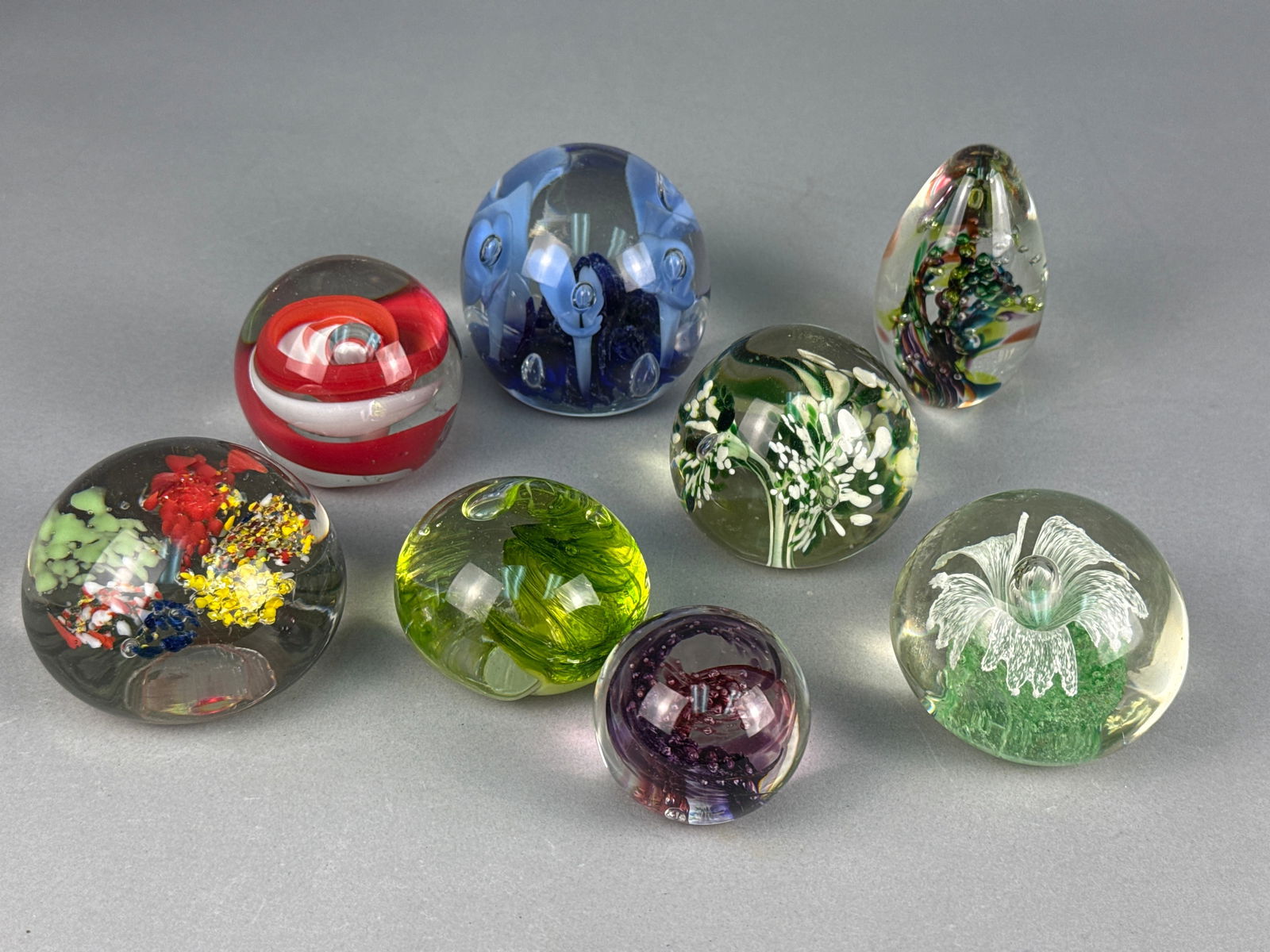 A Collection of Eight Assorted Modern Art Glass Paperweights: This lot comprises eight assorted glass paperweights, primarily spherical with one ovoid form. The paperweights exhibit various internal decorative styles including abstract patterns, controlled bubbl