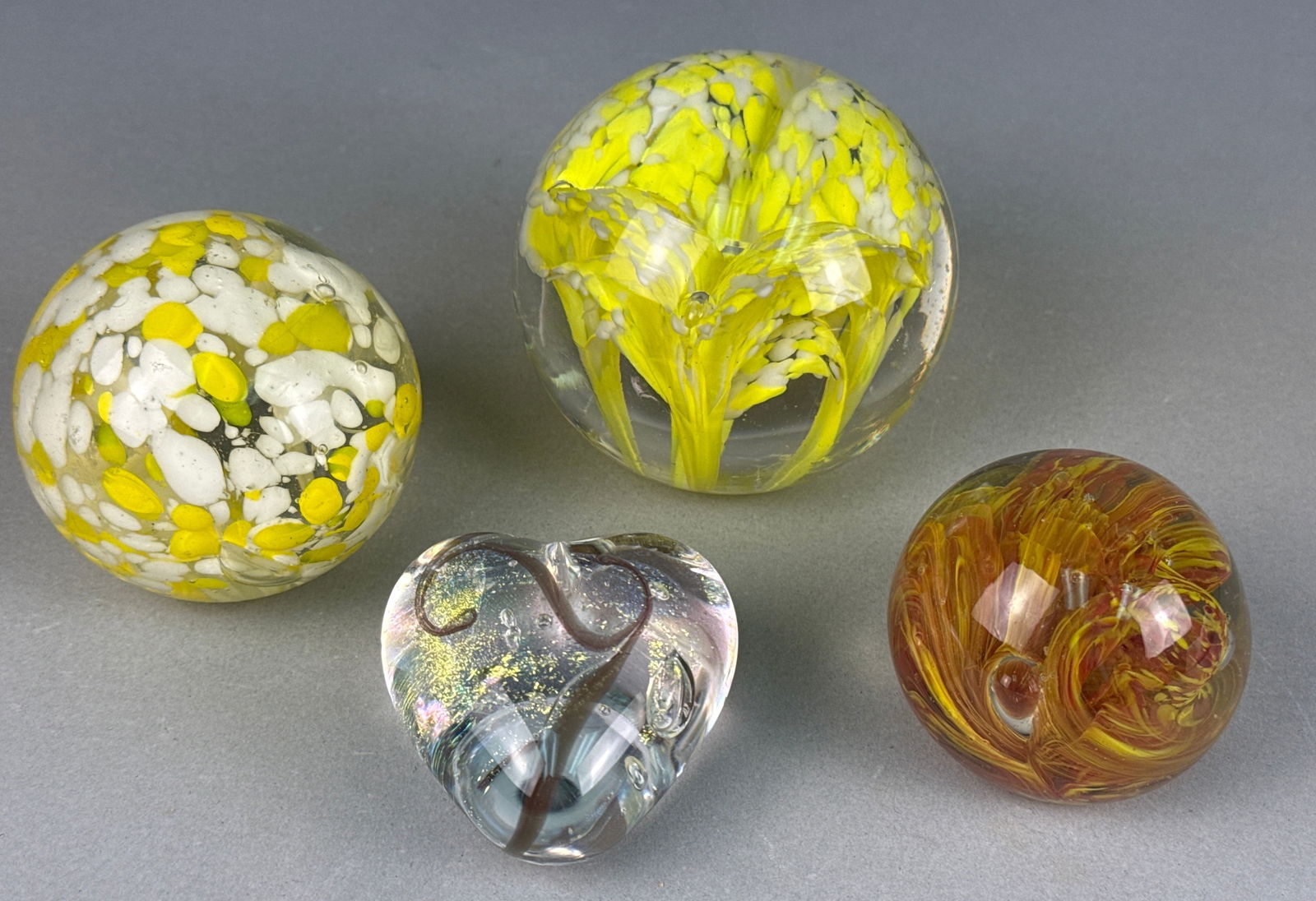 A Collection of Four Modern Art Glass Paperweights, Including a Robert Held Heart and a 'Made in: A collection of four modern art glass decorative objects, comprising three spherical paperweights and one heart-shaped paperweight, all crafted from clear glass with internal colored designs. The hear