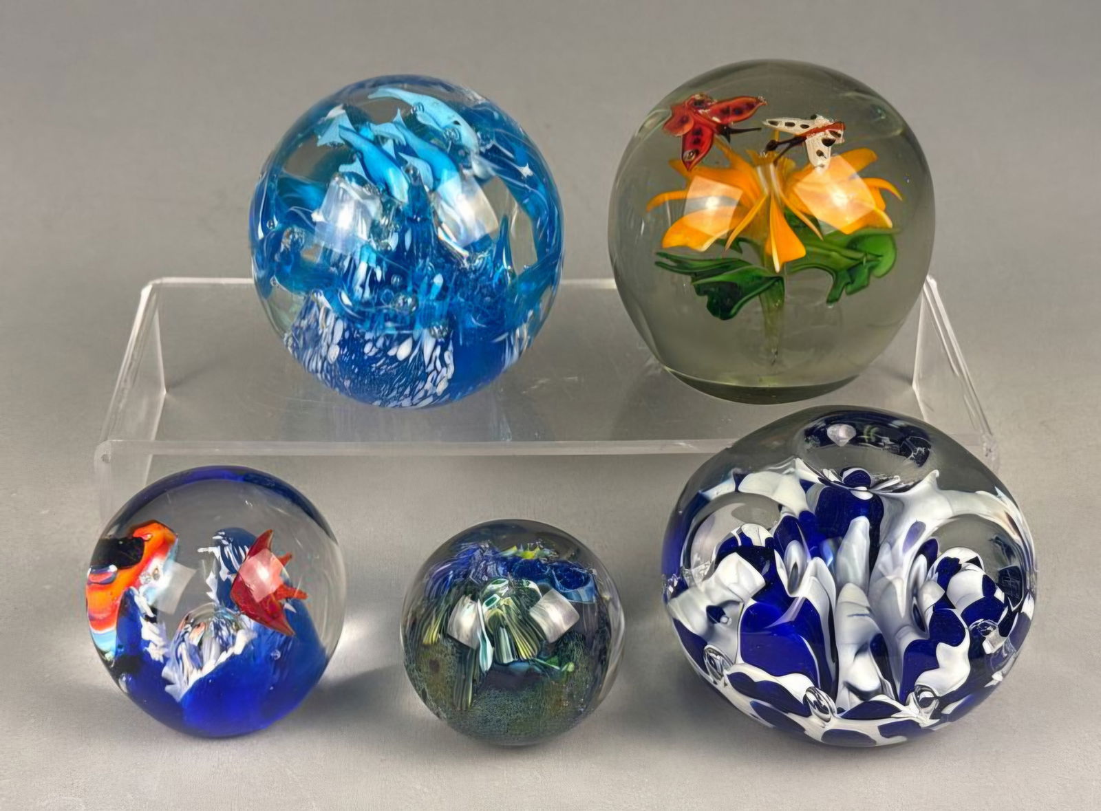 A Collection of Five Art Glass Paperweights, one marked 'HAND MADE BY ST. CLAIR': This lot comprises five spherical art glass paperweights, each featuring unique internal lampwork designs. The largest paperweight on the top left contains blue and white swirls with light blue dolphi