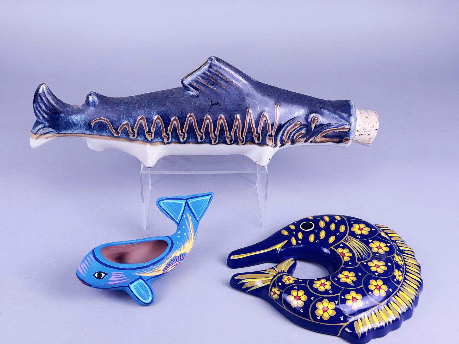 A Group of Three Mexican Folk Art Hand-Painted Ceramic Fish and Whale Figures: A group of three Mexican folk art hand-painted ceramic figures. The largest is an approximately 14-inch long glazed pottery bottle or flask, styled as a dark blue fish or shark, featuring a mottled, t