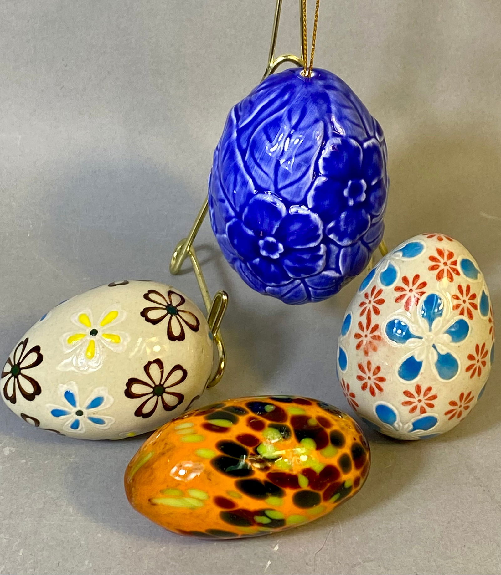 A Collection of 4 Decorative Ceramic and Art Glass Easter Eggs: The collection consists of four decorative eggs utilizing diverse materials and styles primarily focused on spring or Easter themes. The group includes one glossy cobalt blue ceramic or porcelain hang