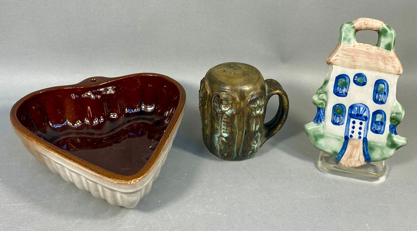 A Trio of Mixed Ceramic Wares including an M A Hadley Country Scene Blue Wall Plaque: A collection of three distinct pieces of ceramic ware. The lot includes a hand-painted ceramic wall plaque, a handled ceramic shaker, and a stoneware baking mold. The first item is a flat-backed cera