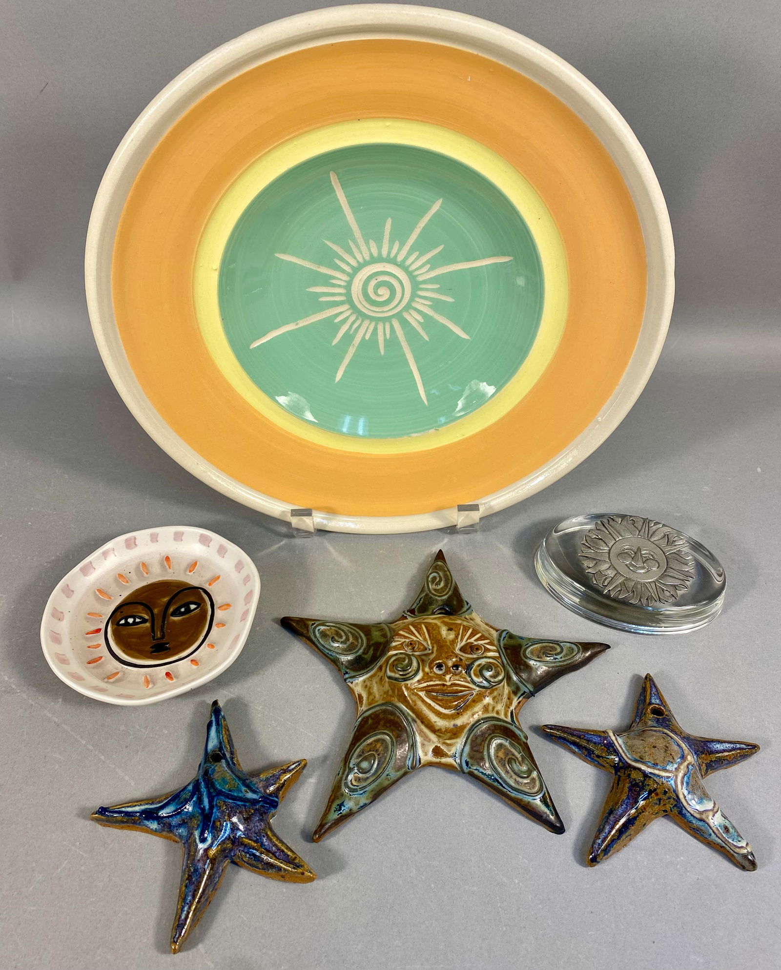 A Collection of Six Decorative Ceramic and Glass Objects with Sun and Star Iconography: The collection comprises six decorative objects centered on celestial iconography, primarily utilizing sun and star motifs. The primary piece is a large circular glazed earthenware charger featuring c