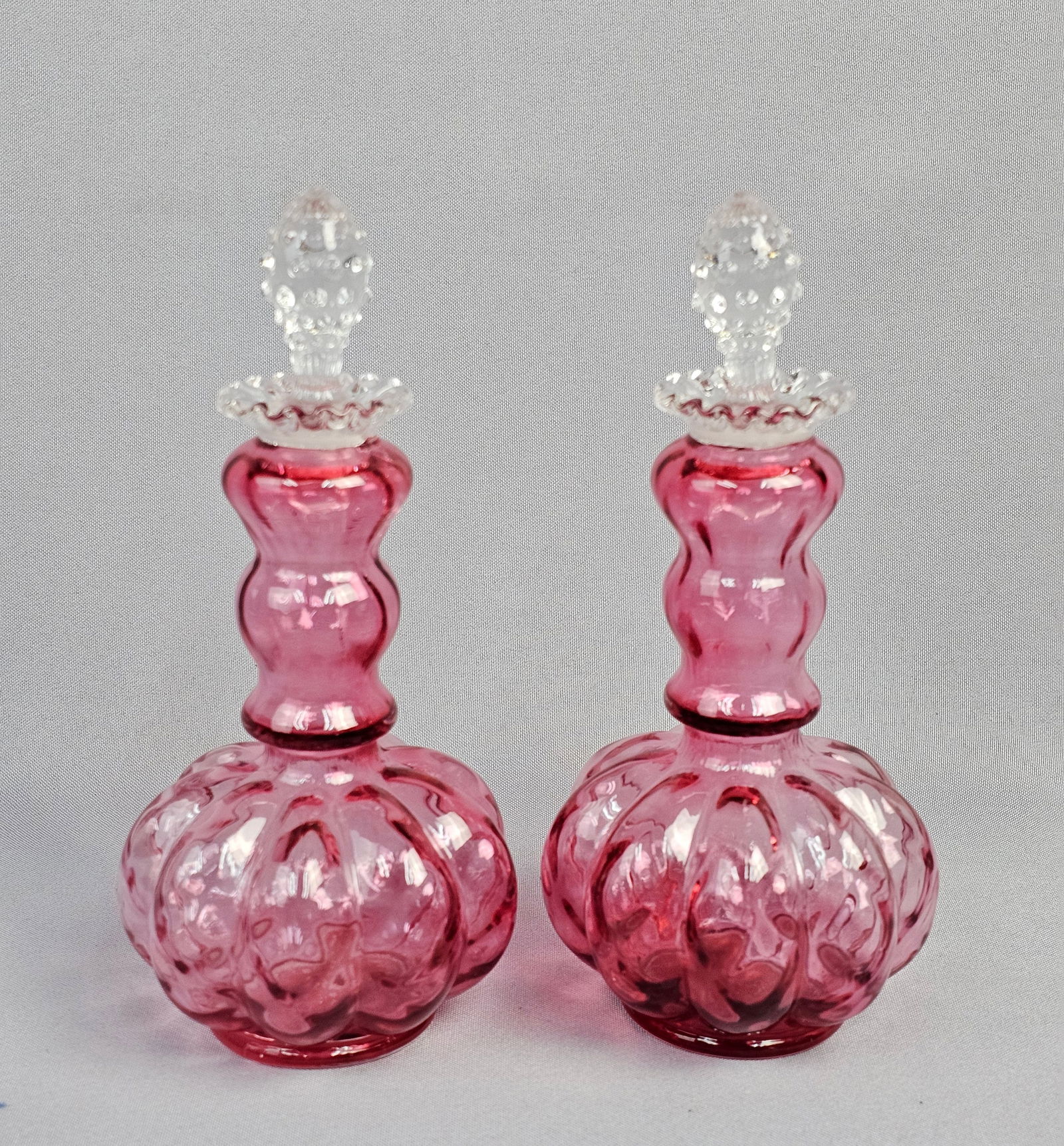 A Pair of Fenton Cranberry Diamond Optic Melon Glass Perfume Bottles: A pair of Fenton Cranberry Diamond Optic Melon Glass Perfume Bottles. Each bottle is crafted from cranberry or deep pink colored glass, featuring a bulbous, vertically ribbed lower body that transitio