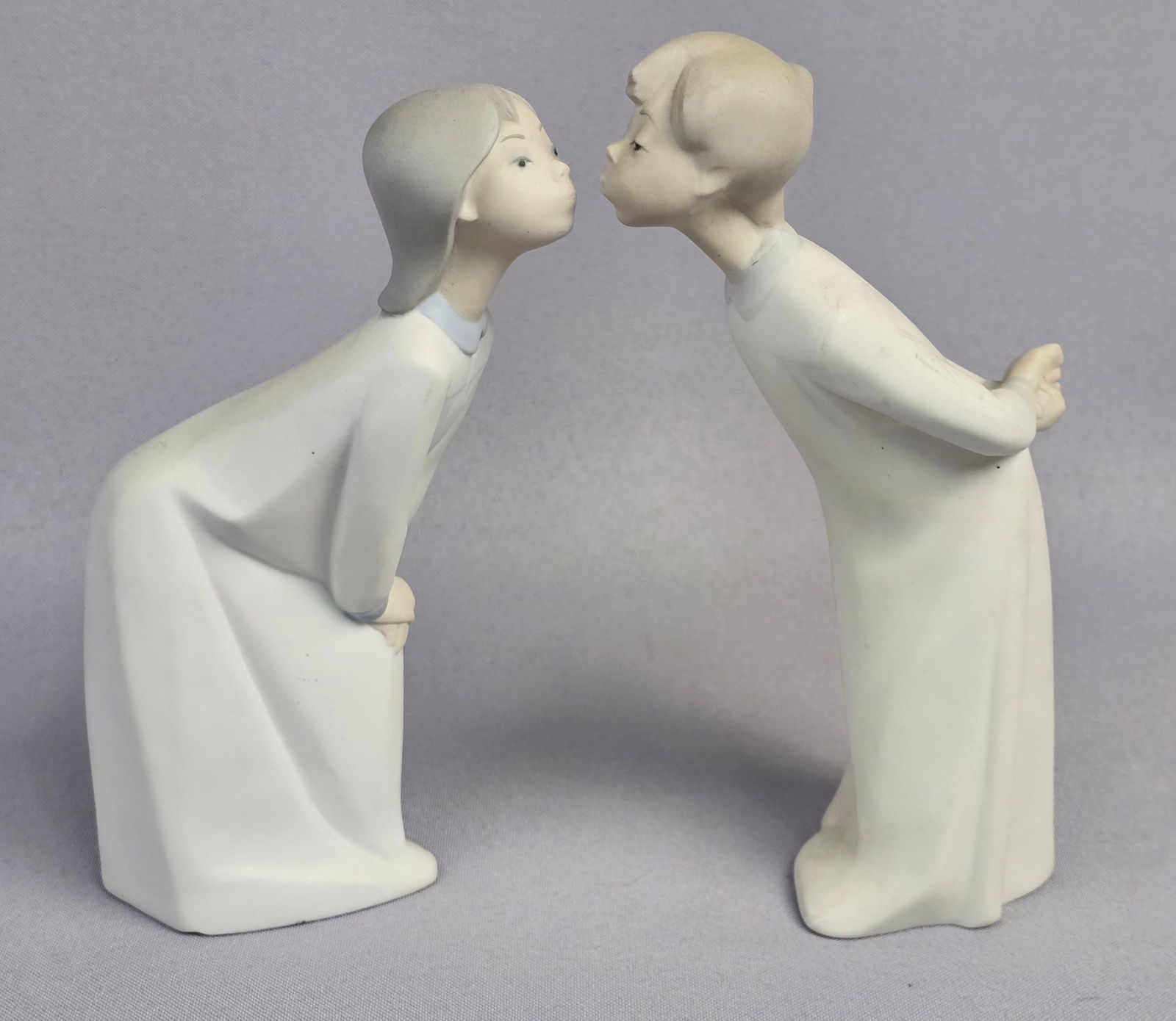 A Pair of Lladro Blowing Kisses Boy and Girl Figurines: A pair of Spanish porcelain figurines manufactured by Lladro, commonly known as the Blowing Kisses or Kiss Me Goodnight models. The figures depict a boy and a girl, both rendered in white matte-finish