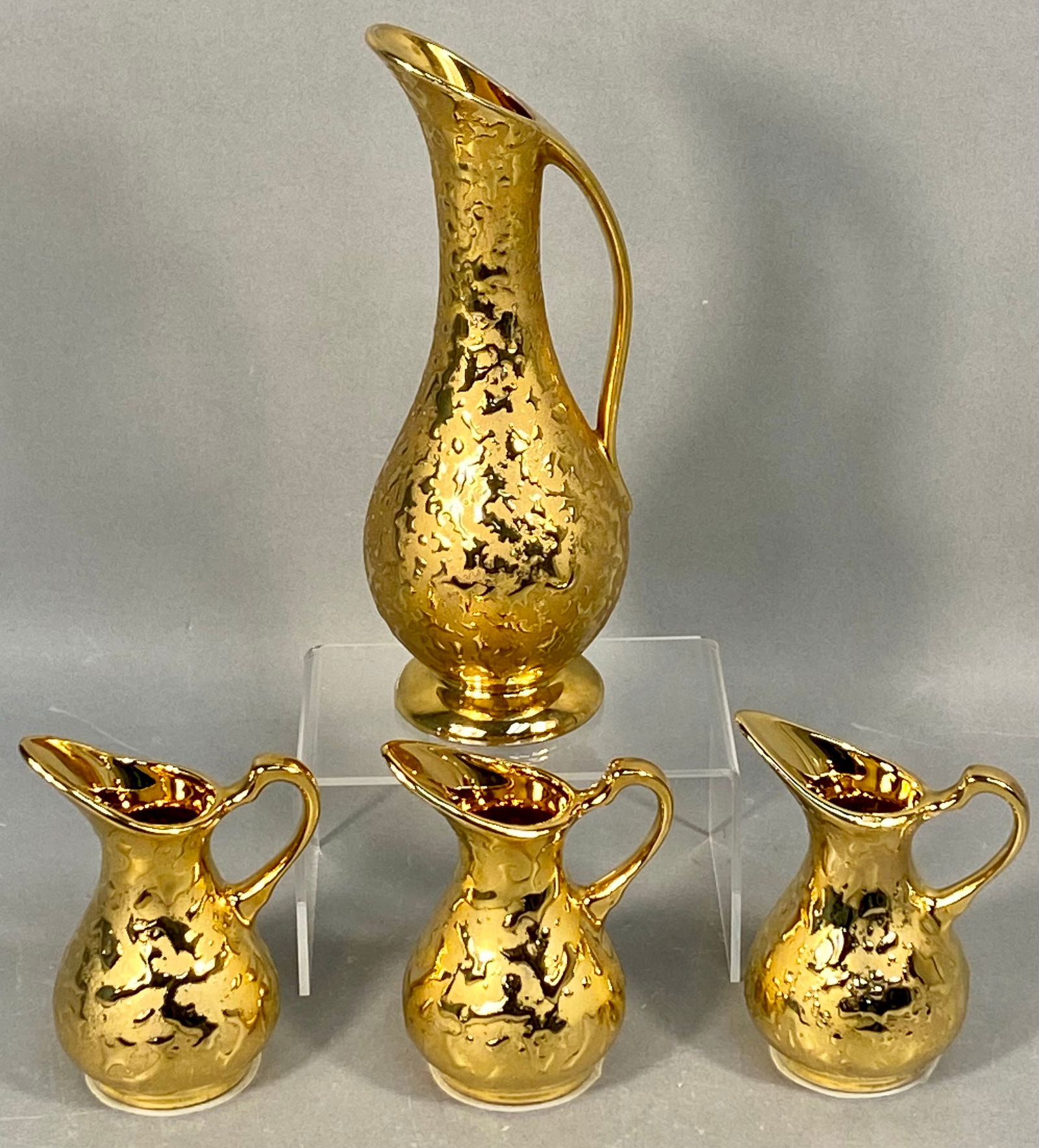 A Collection of Four Dixon Art Studios Weeping Bright 22-KT Gold Ewers and Creamers: A collection of four ceramic vessels attributed to Dixon Art Studios, featuring the distinctive Weeping Bright Gold finish. The set consists of one large ewer or pitcher and three smaller miniature cr
