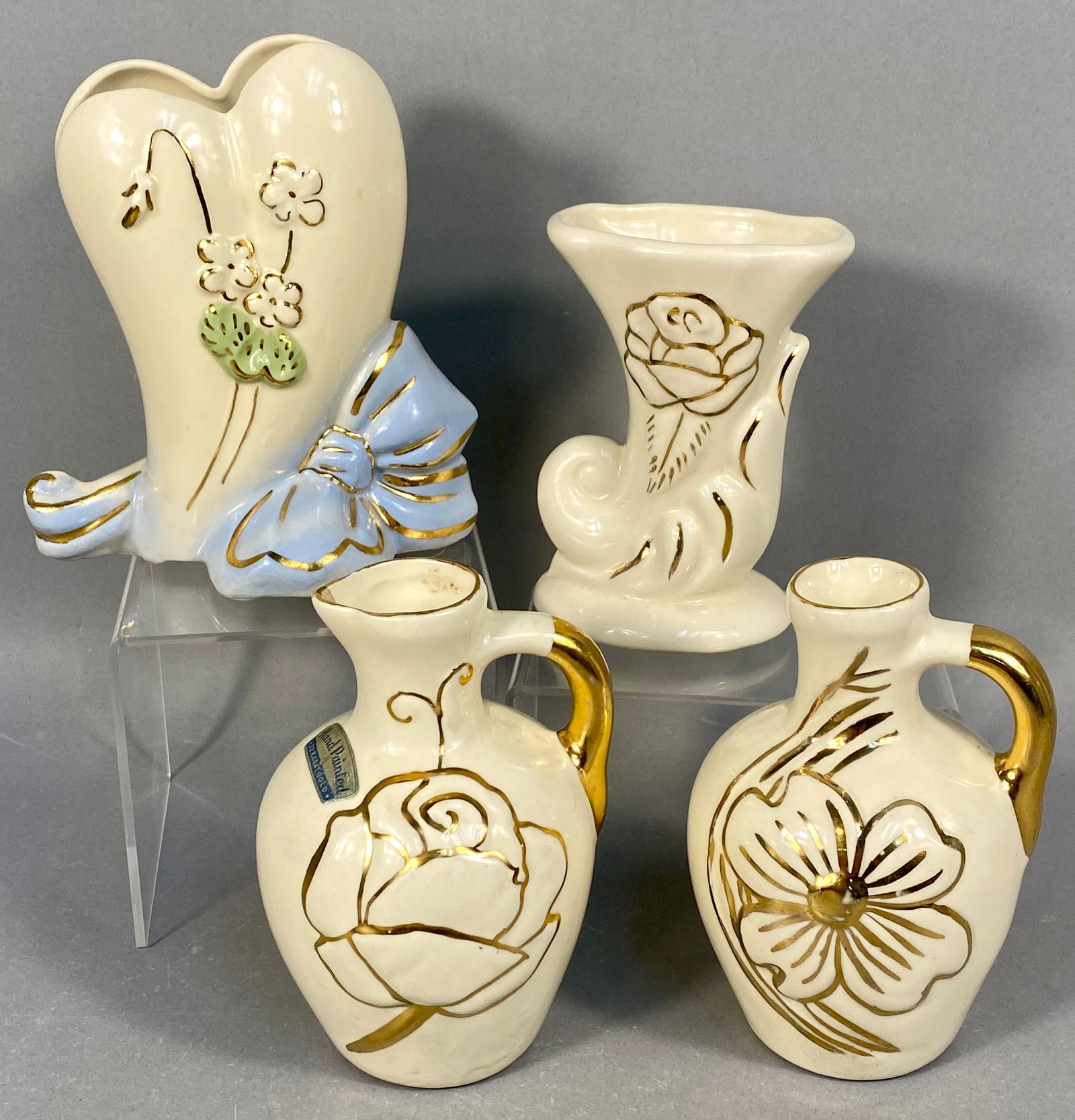 A Collection of Four Mid-Century Cream Ceramic Vessels with 22 Karat Gold Trim: A collection of four decorative novelty vessels, likely dating to the Mid-20th Century, executed in cream-colored glazed ceramic pottery. The items are extensively decorated with hand-painted gold lus