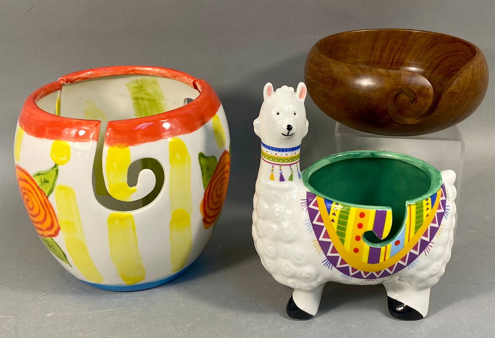 A Trio of Ceramic and Hardwood Yarn Bowls: A trio of yarn bowls designed for knitting or crocheting use, comprising two glazed ceramic vessels and one polished hardwood bowl. The first ceramic bowl is wide and round, decorated with bright, glo