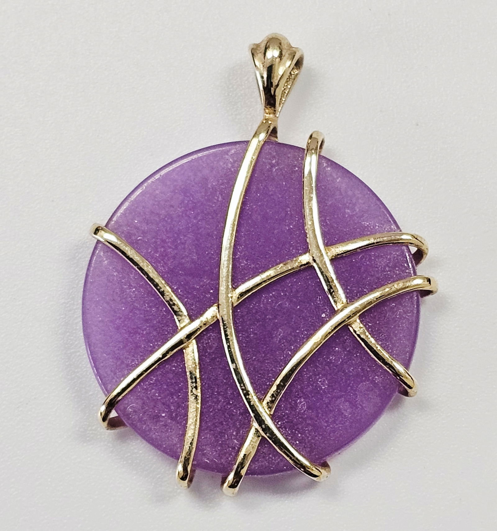 10k Yellow Gold and Purple Stone Wire-Wrapped Pendant: This circular pendant features a flat, polished purple stone set within a 10k yellow gold wire-wrapped mounting. The abstract wire design crosses over the front of the stone in an irregular pattern an