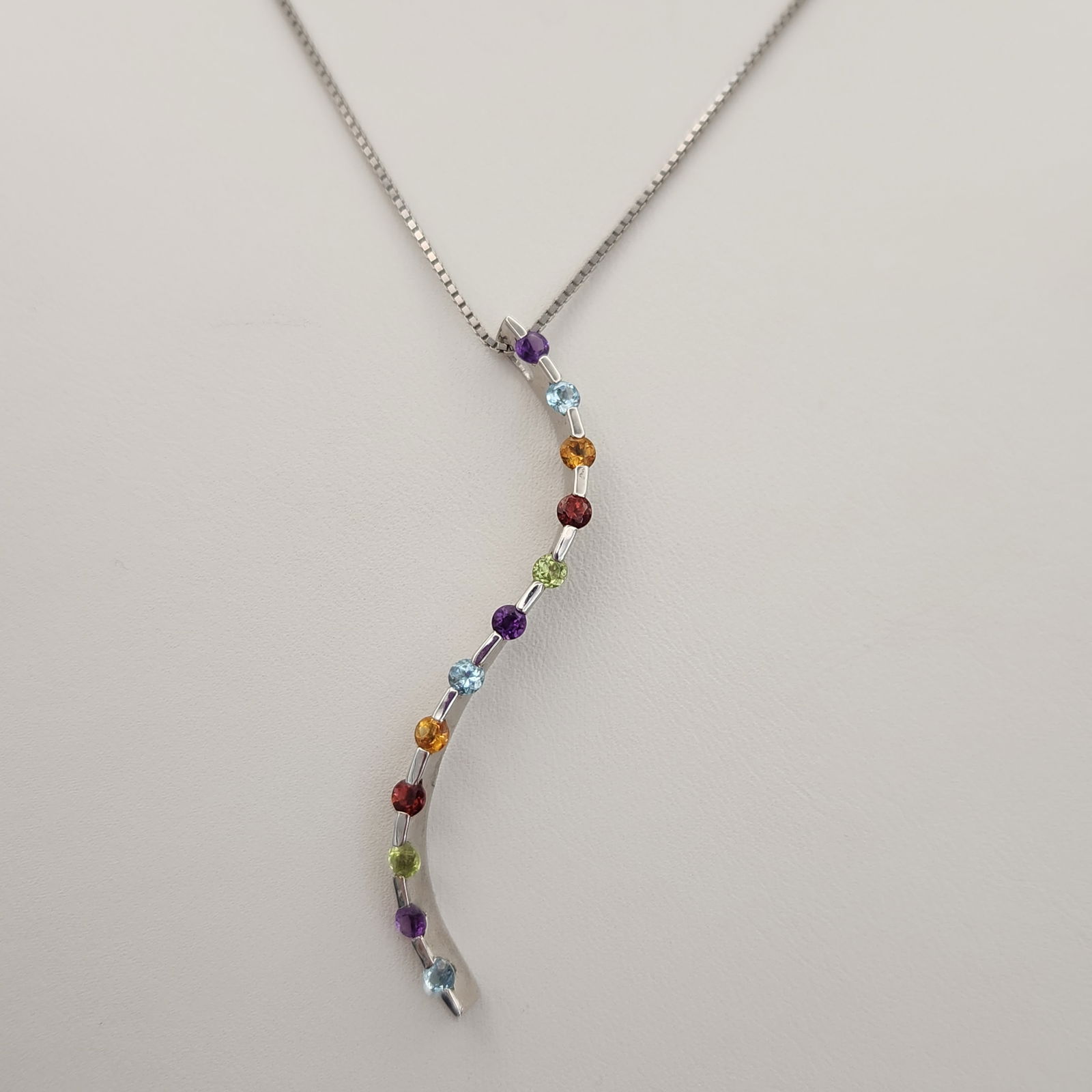 14K Gold Multi-Gemstone Necklace 6.4 Grams: This contemporary necklace is constructed of 14K white gold and features an S-curved pendant set with round-cut multi-colored gemstones. The gemstones include amethyst, blue topaz, citrine, garnet, an
