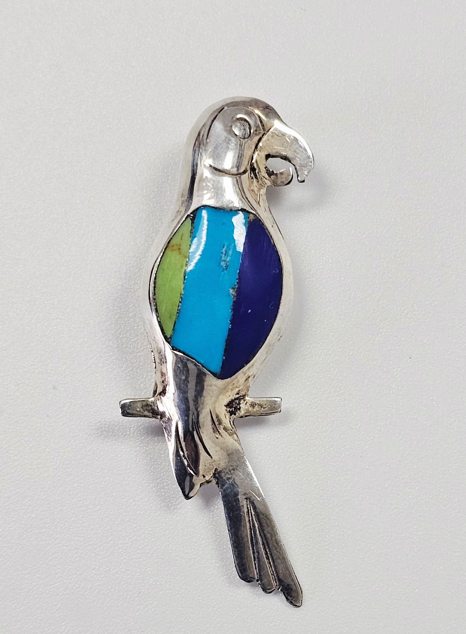 A Mexican Sterling Silver and Multi-Stone Inlay Parrot Brooch: A vintage Mexican sterling silver brooch designed in the form of a stylized parrot perched upon a branch. The bird's body features three vertical panels of inlaid hardstones in shades of green, light