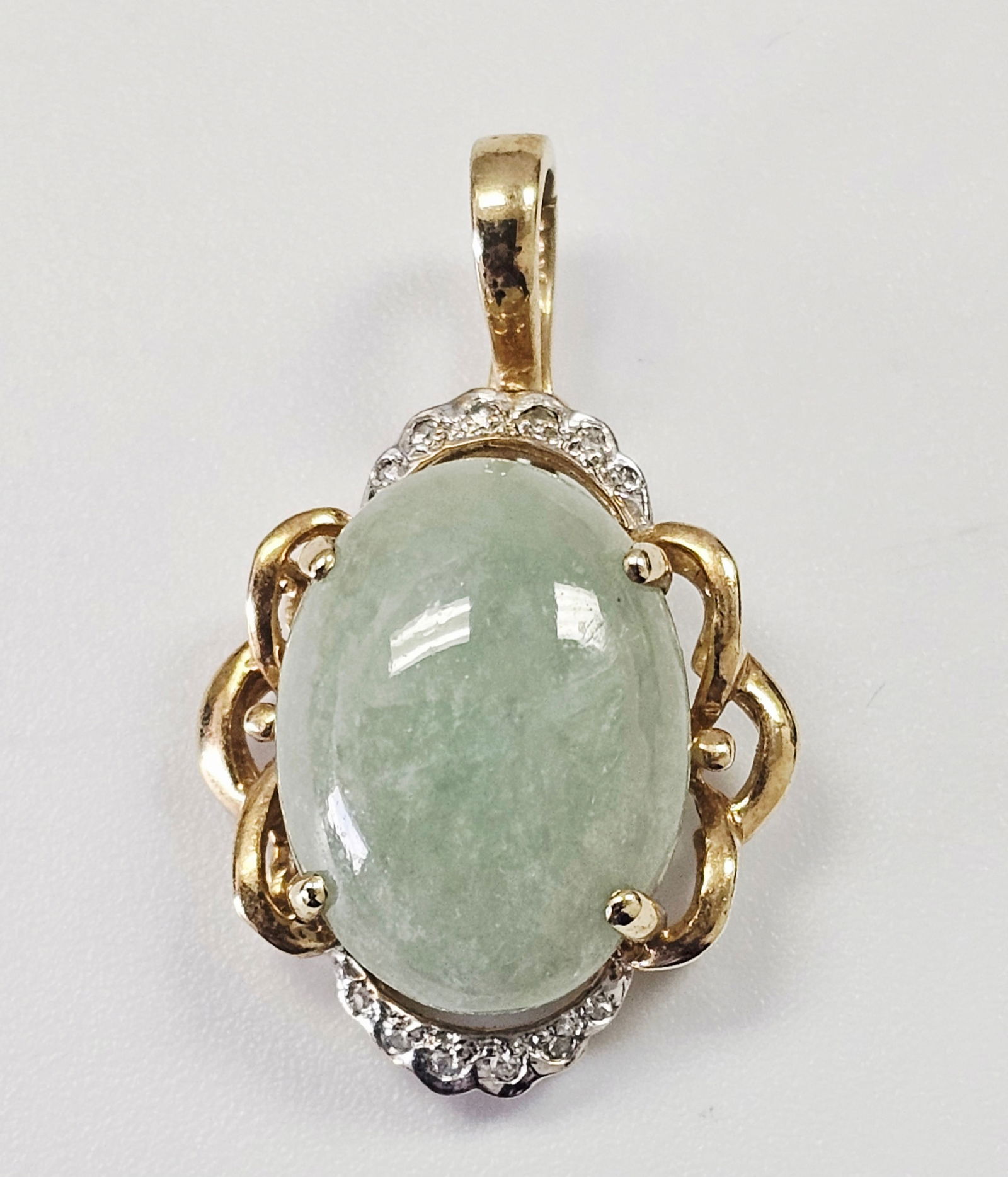 A 10k Yellow Gold and Pale Green Cabochon Stone Pendant with Diamond Accents: A 10k yellow gold pendant featuring a centrally set, oval cabochon-cut pale green stone. The stone is secured by four prongs and surrounded by an ornate, openwork frame with scrolled motifs. The setti