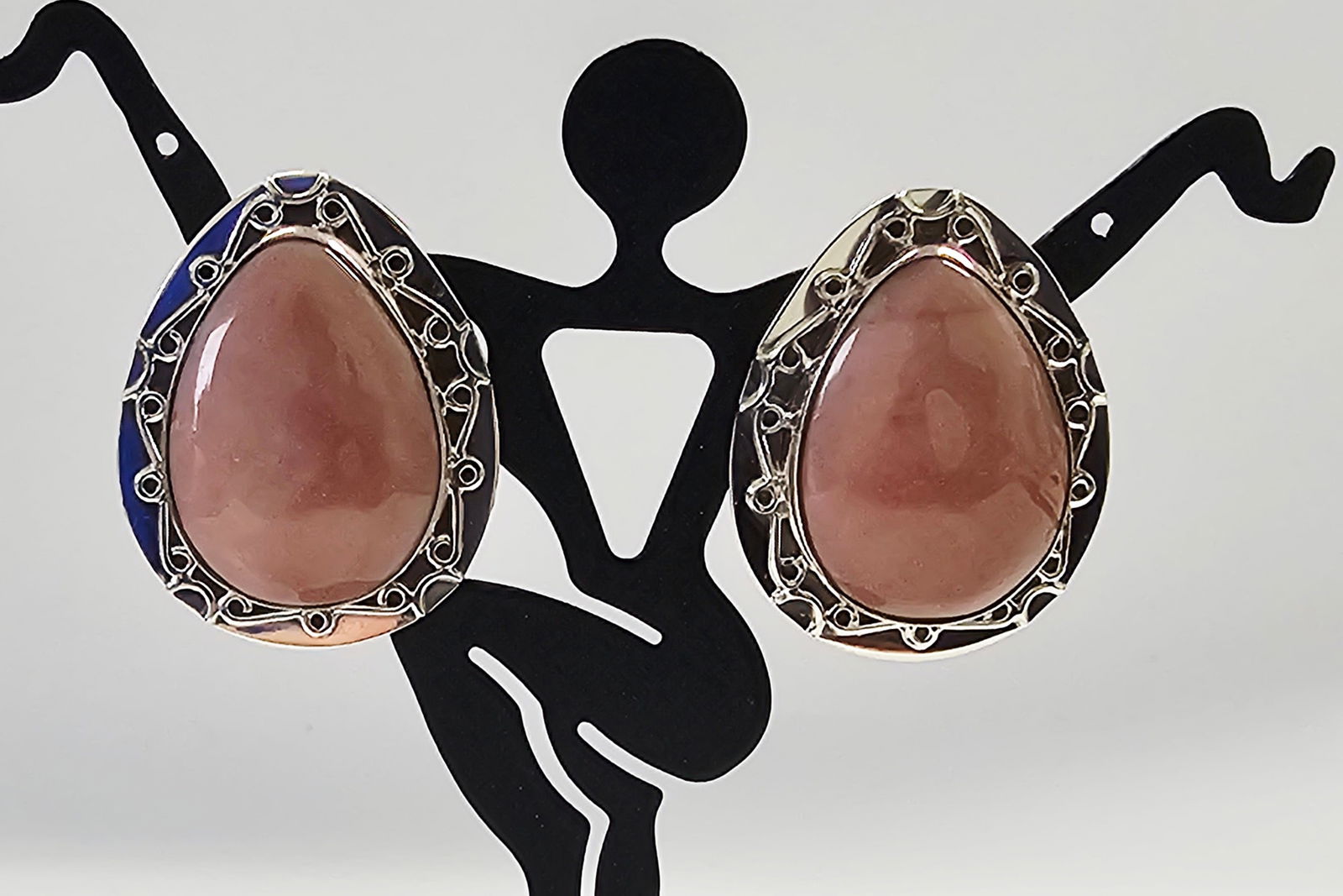 A Pair of Sterling Silver and Rose-Colored Stone Cabochon Post-Back Earrings: A pair of sterling silver post-back earrings. Each earring features a pear-shaped, dusty rose-colored cabochon stone. The stones are secured in a bezel setting surrounded by an ornate openwork silver