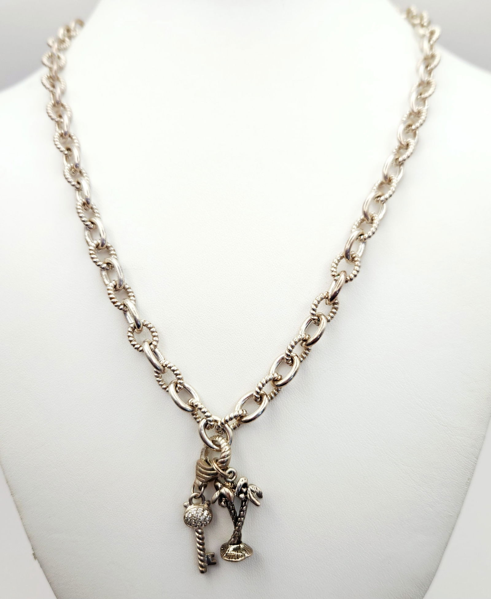 Judith Ripka Sterling CZ Necklace 56.0 Grams: A Judith Ripka sterling silver charm necklace composed of alternating smooth and rope-textured oval links. The design incorporates three charms: a palm tree, a heart-shaped lock, and a key featuring