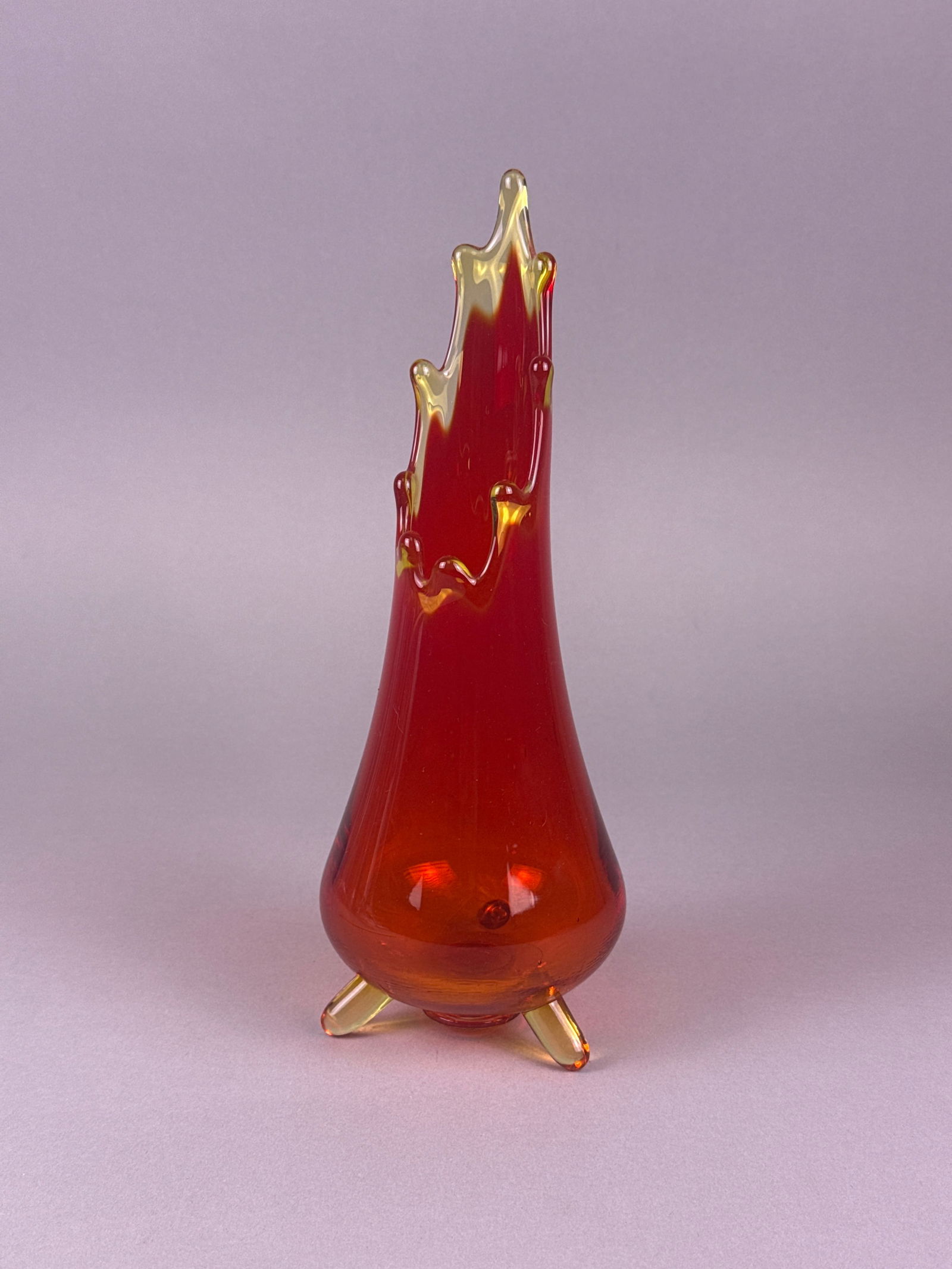 Mid-Century Modern Three-Footed Amberina Flame Swung Vase: The hand-blown vessel exhibits the characteristic Amberina coloring, transitioning dramatically from deep ruby red/cranberry at the high, asymmetrical rim to dense orange/amber toward the bulbous base