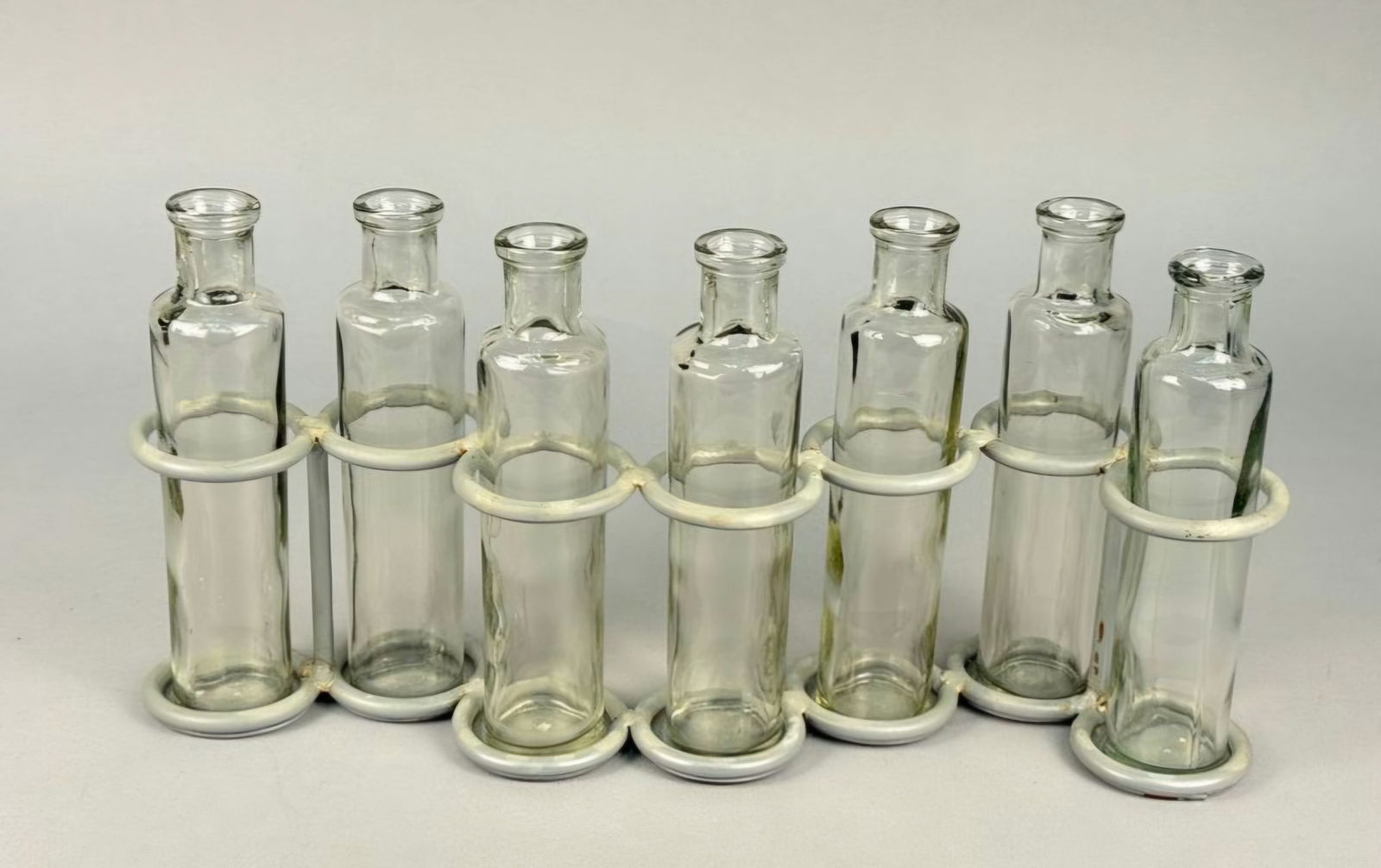 A Collection of Eight Vintage Glass Propagation Bottles and Metal Stand: A collection comprising eight cylindrical, narrow-necked glass bottles and a single continuous metal wire rack designed for use as a propagation station or vase array. The glass exhibits a clear or pa