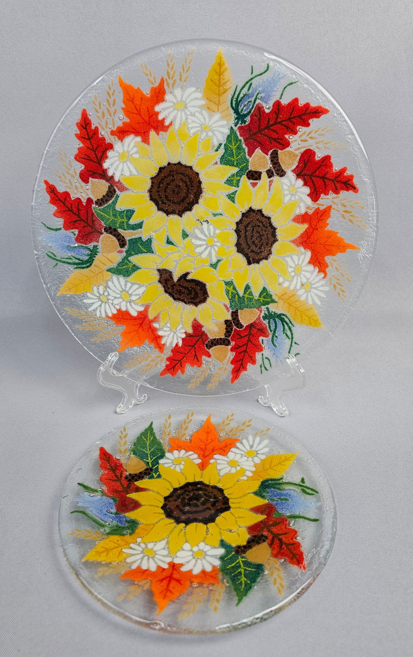 A Pair of Peggy Karr Signed Fused Glass Autumn Sunflower Plates: A pair of contemporary fused art glass items attributed to Peggy Karr, featuring the Autumn Sunflower motif. The lot consists of one large round decorative plate or charger and one smaller matching ci