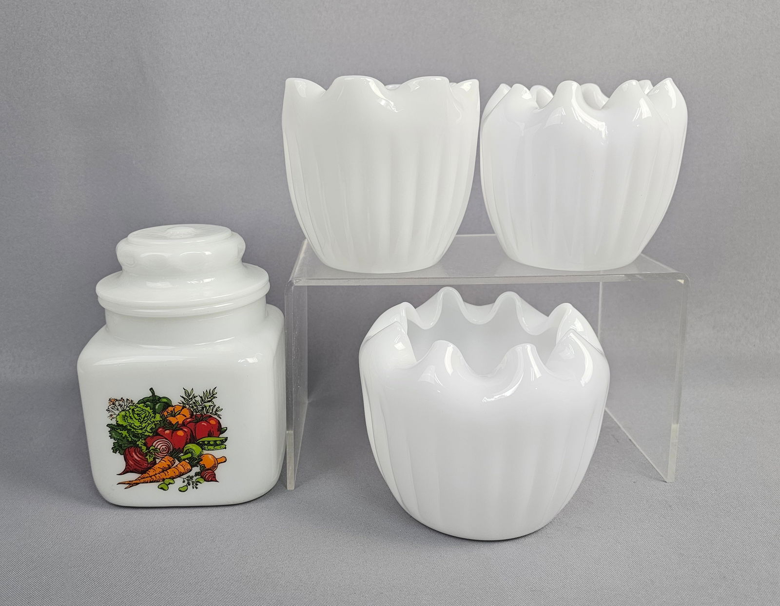 A Collection of Four Milk Glass Pieces Ruffled Rose Bowls and Lidded Kitchen Jar: A collection of four pieces of opaque white Milk Glass dating to the Mid-Century period. The collection includes three matched decorative vessels/vases and one storage jar with a lid. Square-bodied st