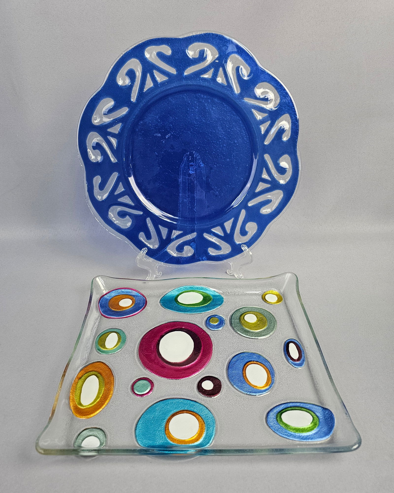 A Pair of Modern Decorative Glass Serving Pieces Inc Lori Siebert for Silvestri Plate: A pair of modern or contemporary decorative glass serving pieces. The first item is a circular charger plate crafted from deep blue glass. The second item is a square serving platter made of clear, te