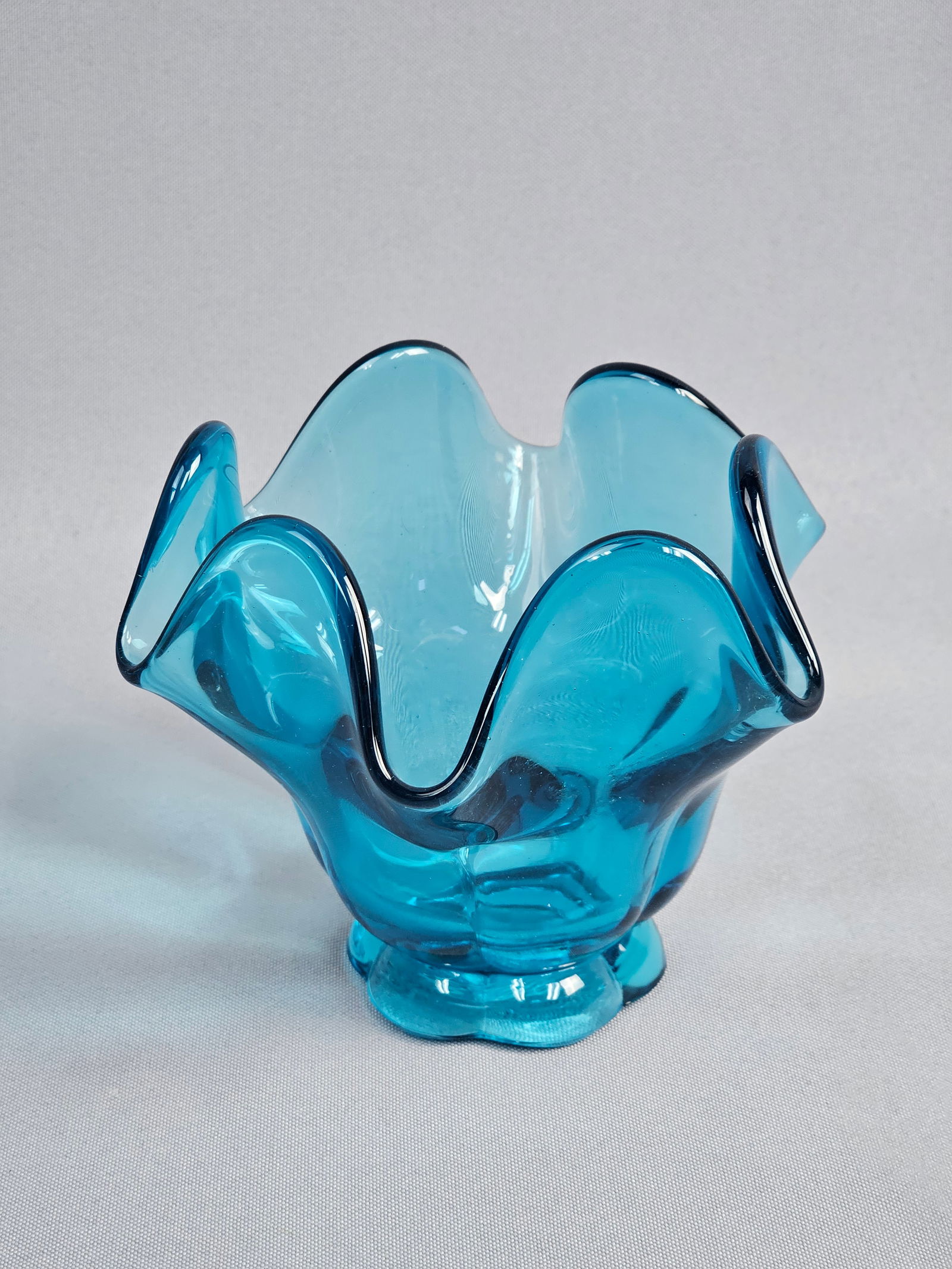 A Mid-Century Modern Style Blue Glass Handkerchief Vase: This mid-century modern style handkerchief vase is composed of translucent blue glass. The vessel features a wide, ruffled rim and is supported by a scalloped, six-lobed foot. There are no apparent ma