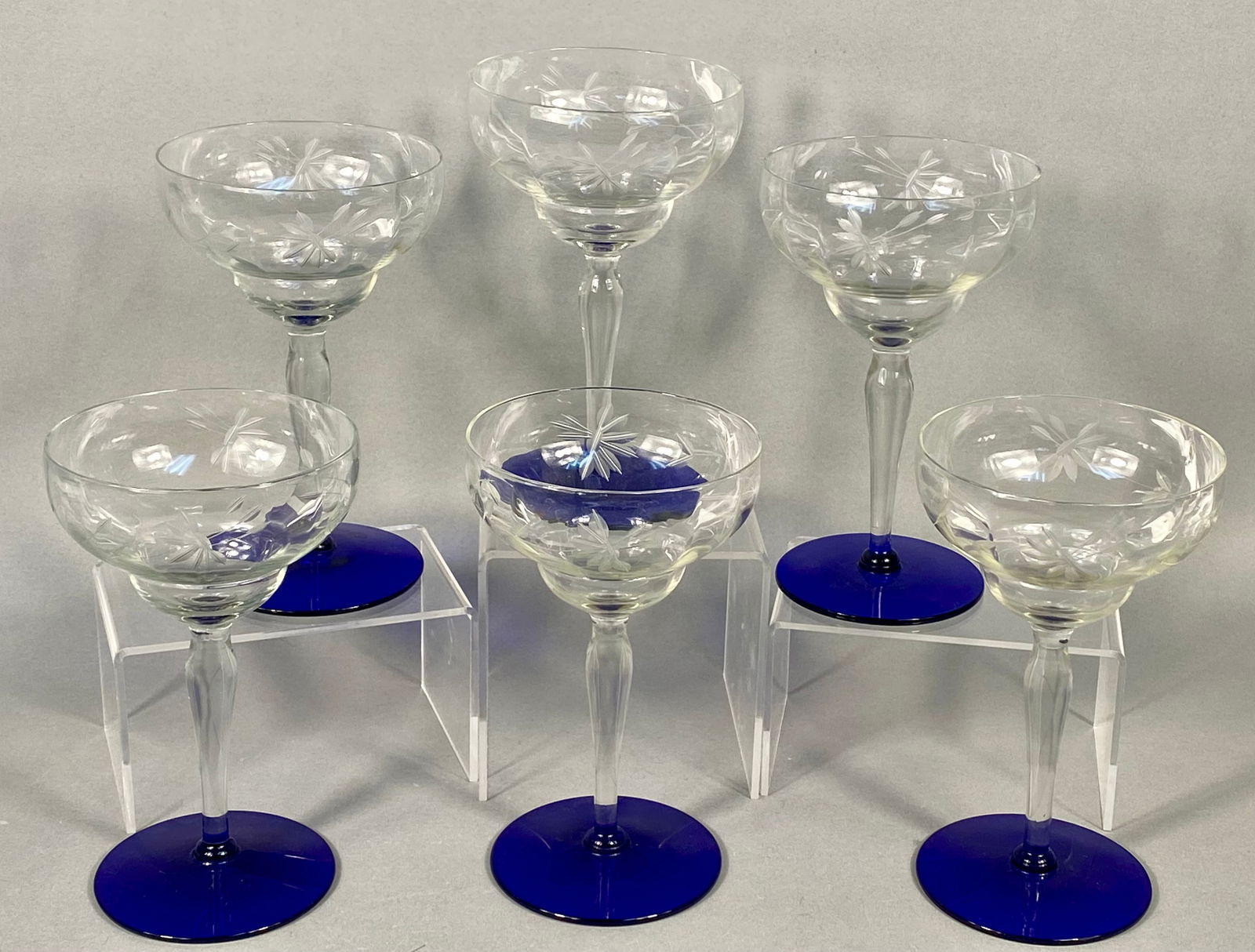 A Set of Louie Weston Etched Crystal and Cobalt Blue Champagne Sherbet Glasses: A set of six crystal champagne sherbet glasses produced by Louie Weston, dating to the 1930s. Each piece features a clear glass bowl decorated with an etched floral and starburst pattern. The bowls ar