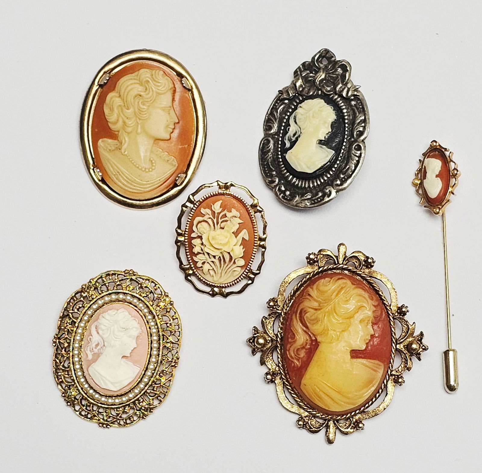 Vintage Costume Jewelry Cameo Brooches and a Stick Pin Inc Avon: A collection of mid-to-late 20th-century costume jewelry cameos in classical revival styles. The group consists of several oval-shaped brooches and a stick pin featuring molded resin or plastic relief