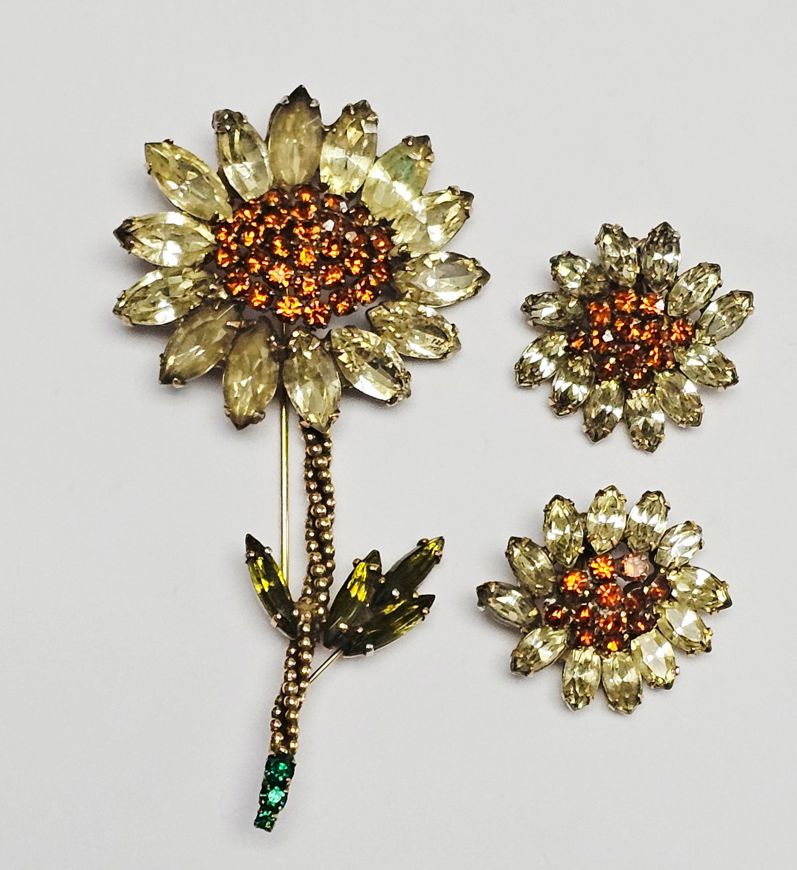 Weiss Rhinestone Sunflower Brooch & Earrings: This collection of vintage costume jewelry by Weiss includes a long-stemmed sunflower brooch and a pair of matching clip-on earrings. The items are crafted from gold-tone metal and feature an array