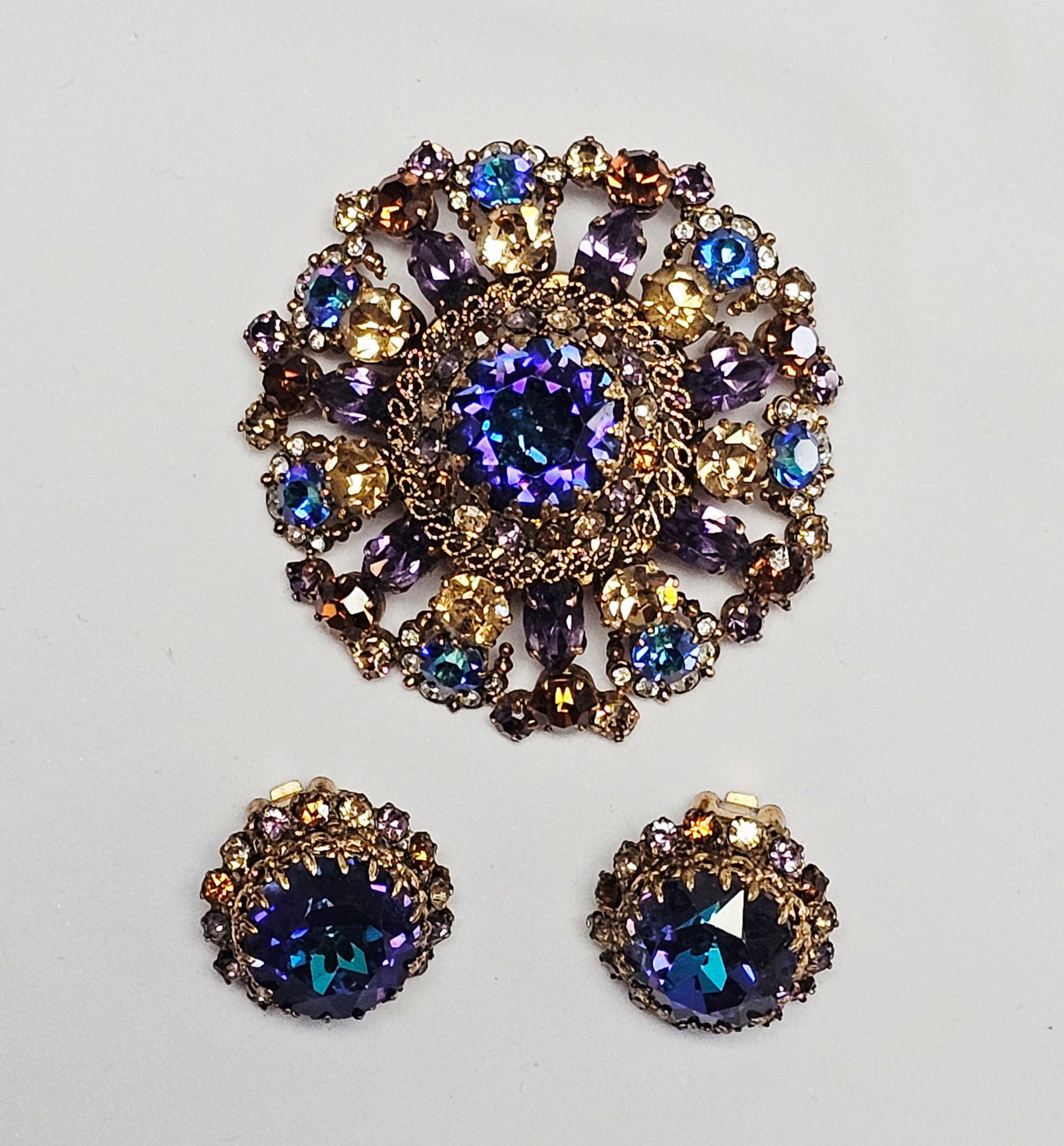 Vintage Austrian Blue and Purple Rhinestone Brooch & Earrings: This trio of costume jewelry consists of a circular brooch and a matching pair of clip-on earrings. Each piece is constructed from gold-tone metal and set with a variety of faceted glass rhinestones.