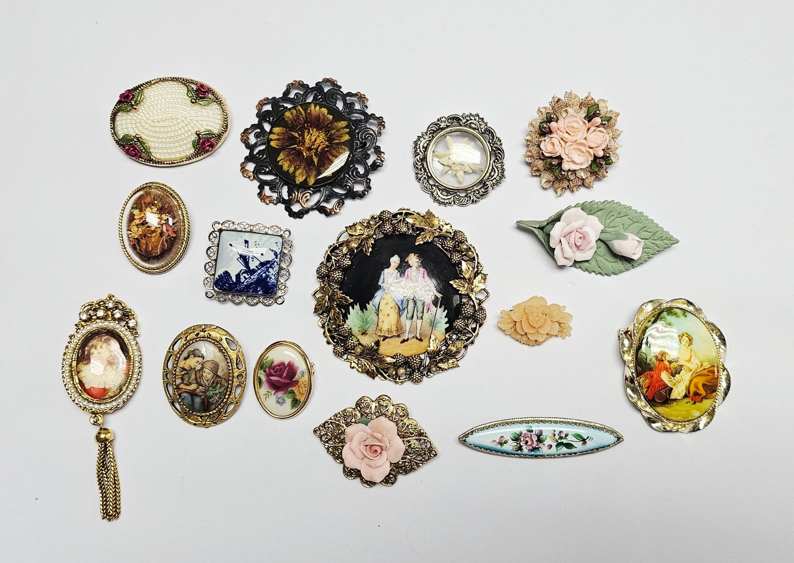 Vintage-Style Costume Jewelry Brooches and Pins: A collection of vintage-style costume jewelry brooches and pins featuring various decorative motifs and techniques. The group includes pieces depicting romantic bucolic scenes, floral designs, a blue