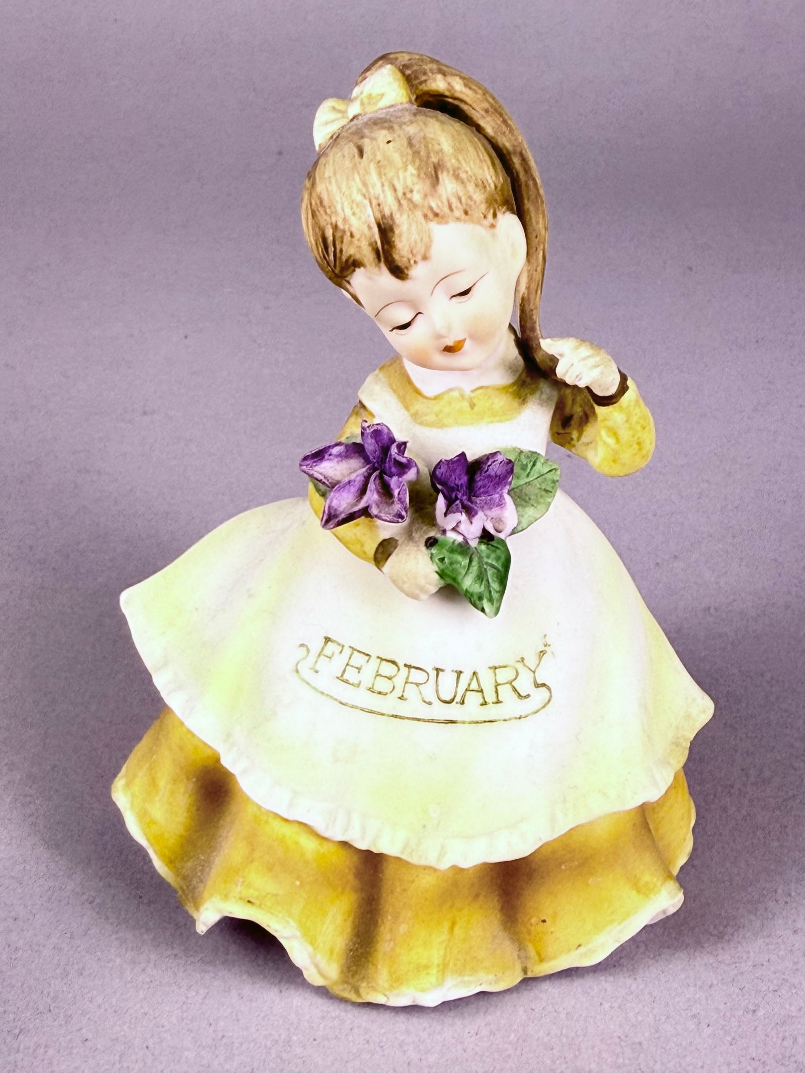 A Pair of Mid-Century Ceramic February Birthday Girl Figurines by Lefton and Josef Originals: Two mid-century ceramic figurines representing the month of February. The first is a bisque porcelain figurine by Geo. Z. Lefton, model KW4200, from the Girl of the Month series. It depicts a girl in