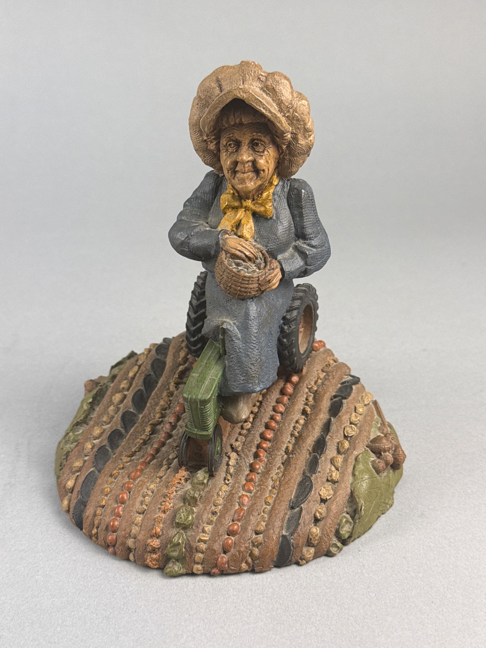 Tom Clark Cairn Studio Gnome Flora Figurine 1989: This hand-painted resin figurine, titled Flora, was created by artist Tom Clark for Cairn Studio in 1989. The piece depicts a gnome-like elderly woman seated on a green tractor. The figure is dressed