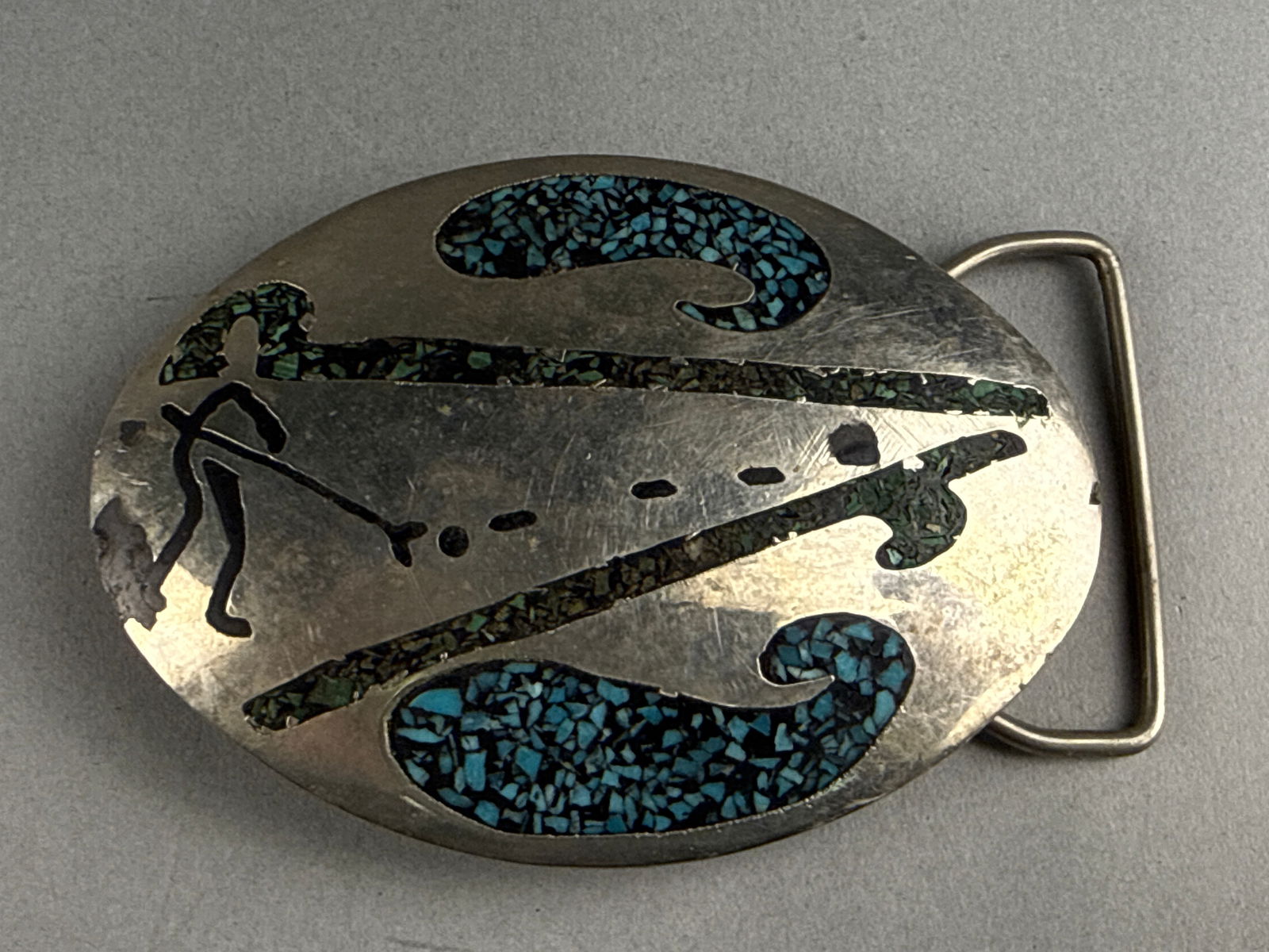Uncle Dunkle's Belts and Buckles Kokopelli Turquoise Chip Inlay Belt Buckle, 1979: An oval silver-toned metal belt buckle produced by Uncle Dunkle's Belts and Buckles, dated 1979. The face features a Southwestern aesthetic depicting a Kokopelli figure and stylized scroll motifs.