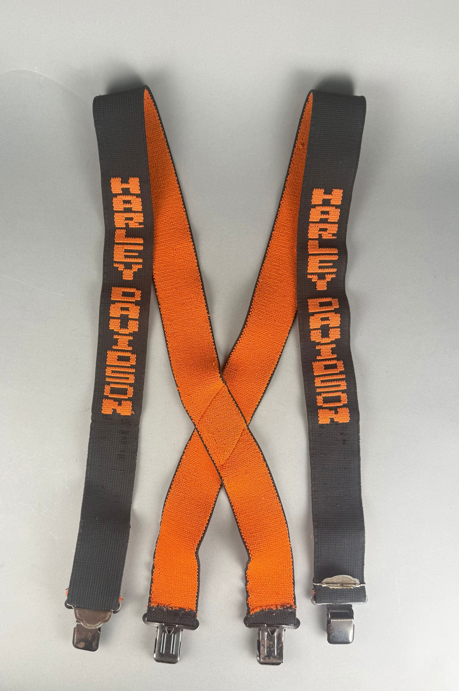 Harley Davidson Woven Logo Elastic Suspenders: A set of Harley Davidson branded suspenders featuring elasticized fabric straps. The straps are designed in black with the Harley Davidson name woven in orange block lettering. The interior of the str